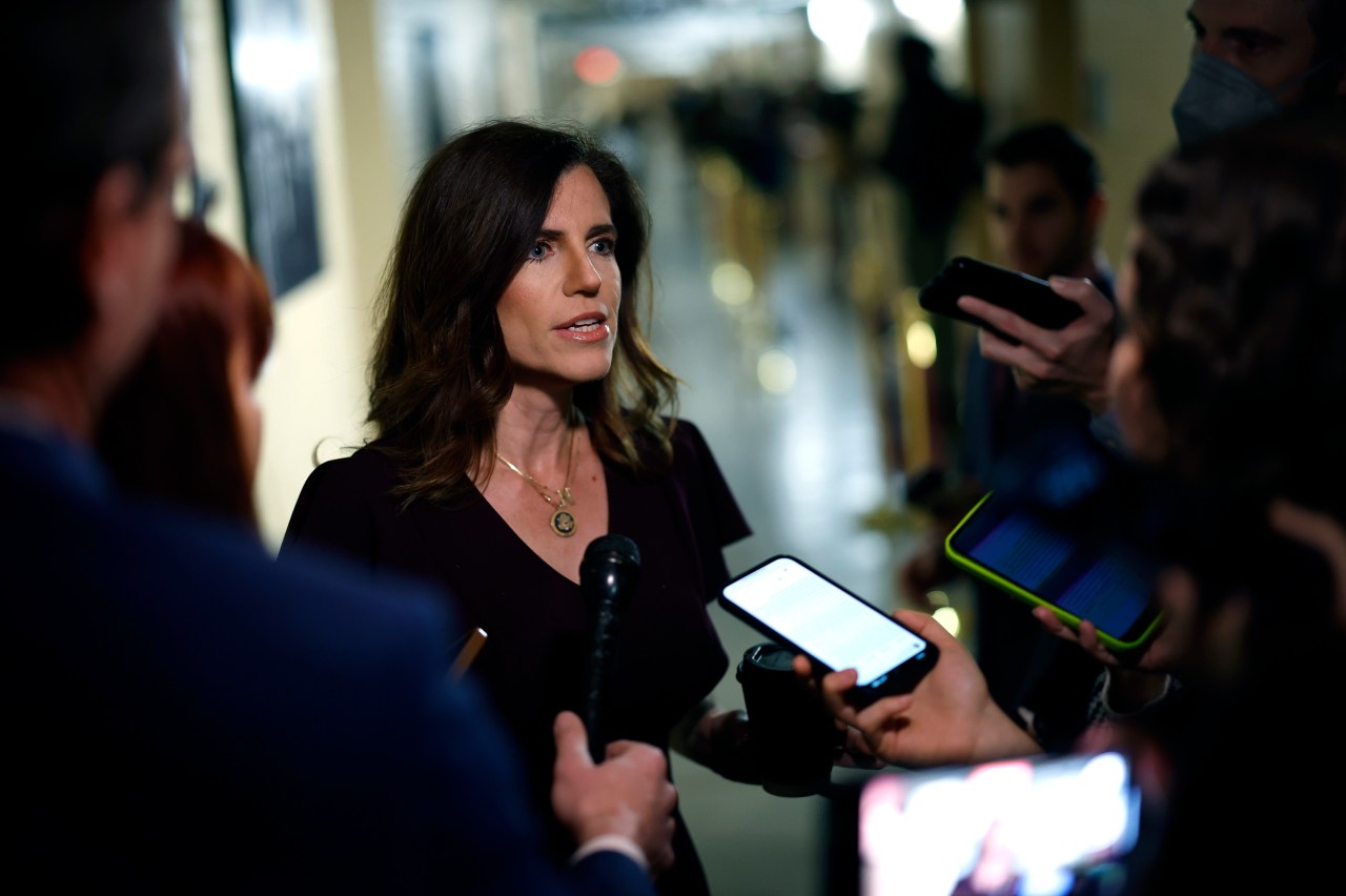 Rep. Nancy Mace stands in a Capitol corridor speaking to reporters holding microphones and phones, wearing a dark burgundy top.