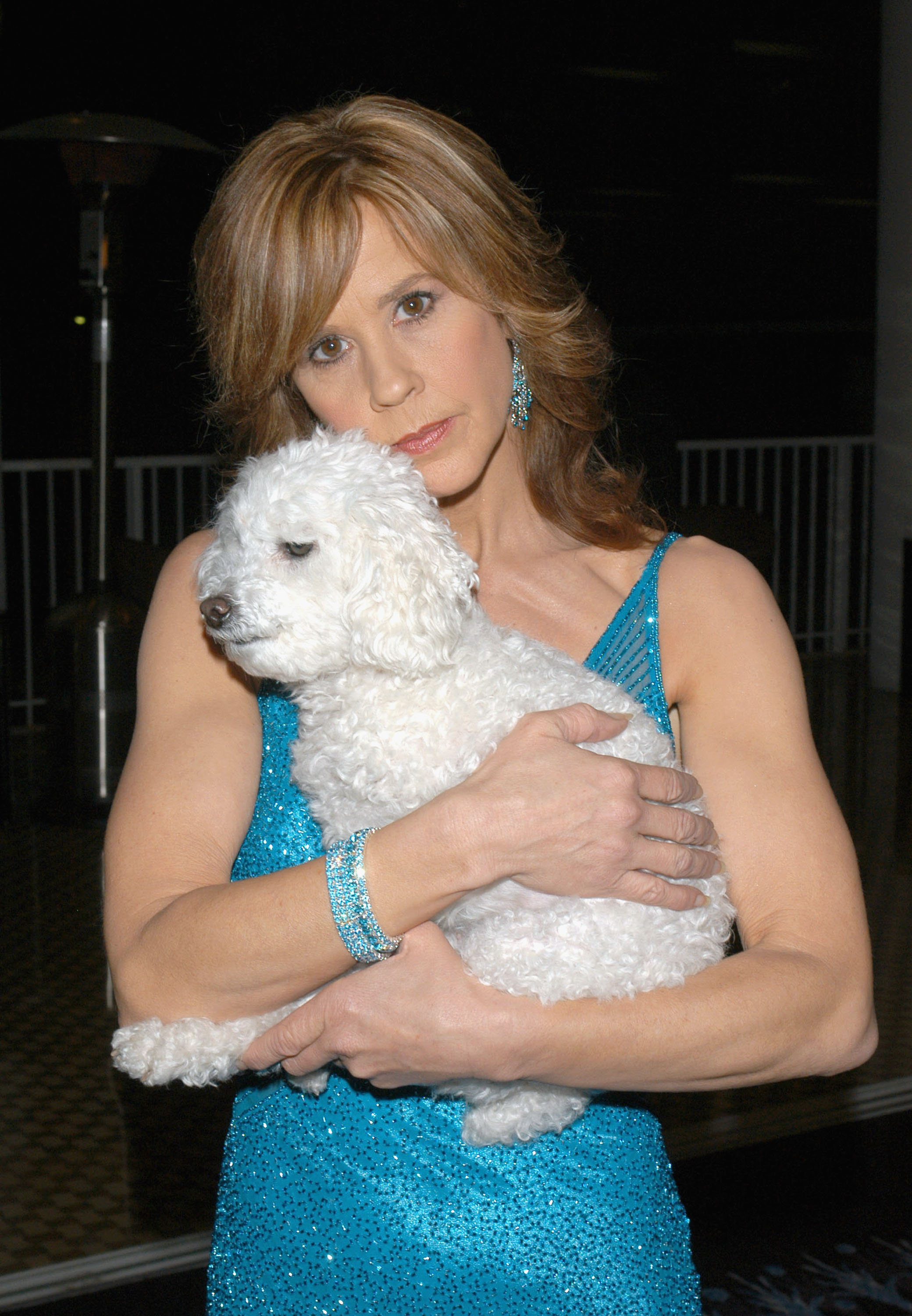 Woman in a blue sequined dress holding a small white fluffy dog close to her chest at an indoor event.