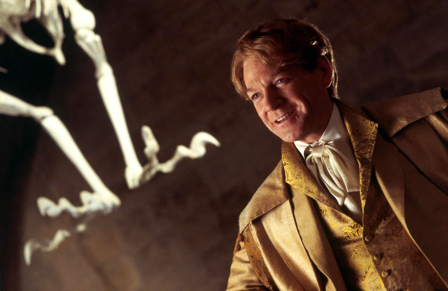 A smiling man in a gold brocade waistcoat and white cravat poses next to a skeleton in a dark, cave-like setting.