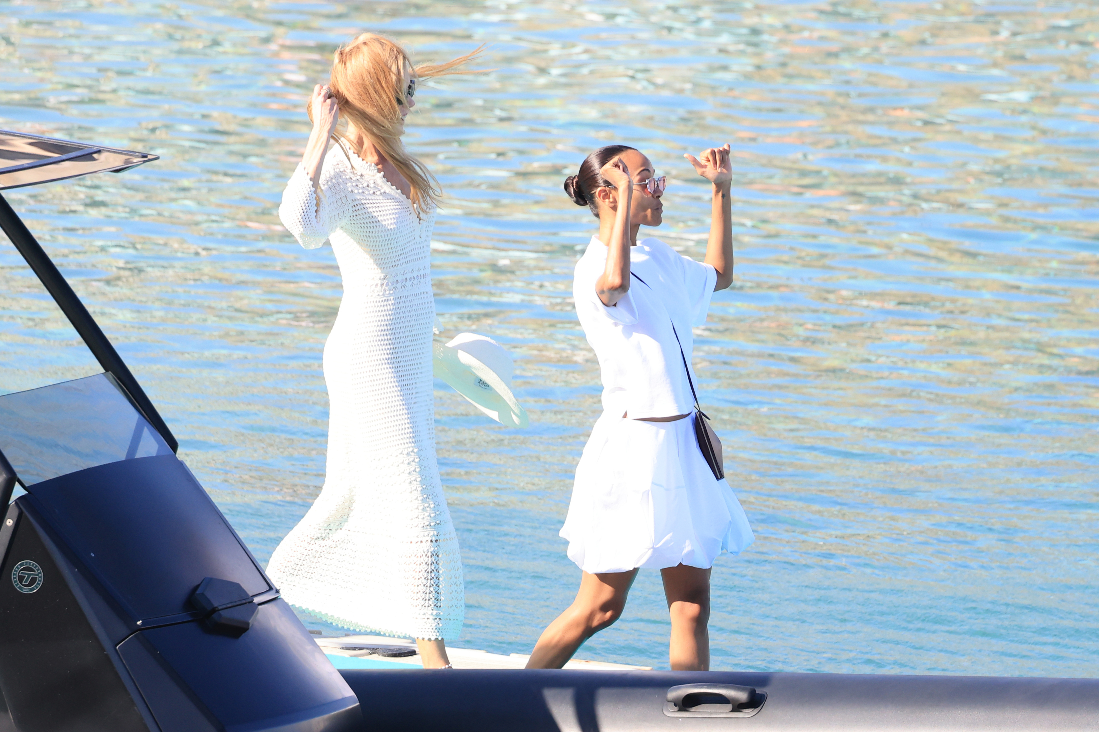 Two women in white outfits standing near a boat with sparkling blue water behind them, one shielding her eyes from the sun.