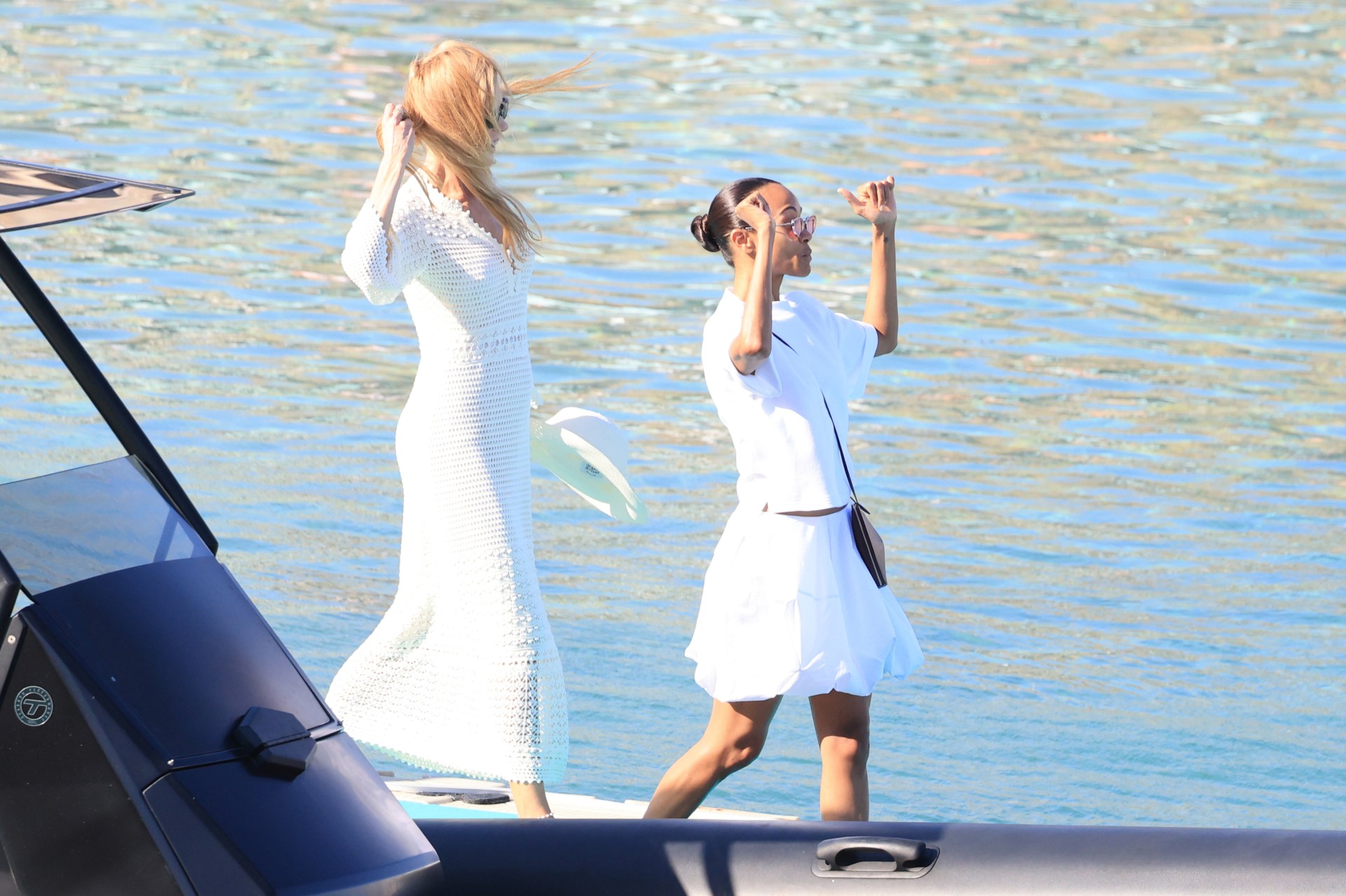 Two women in white outfits standing near a boat with sparkling blue water behind them, one shielding her eyes from the sun.
