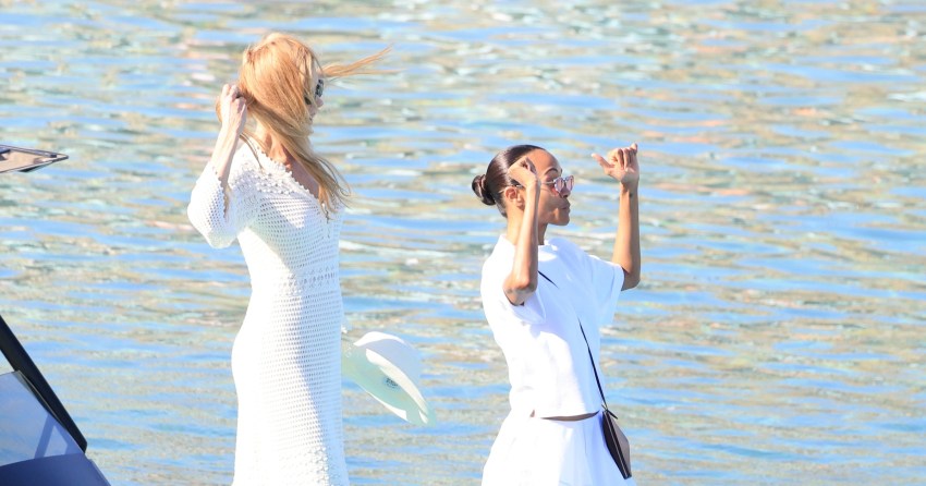 Two women dressed in white outfits stand near a yacht on the water, one with long blonde hair and one with dark hair in a bun, both gesturing animatedly.