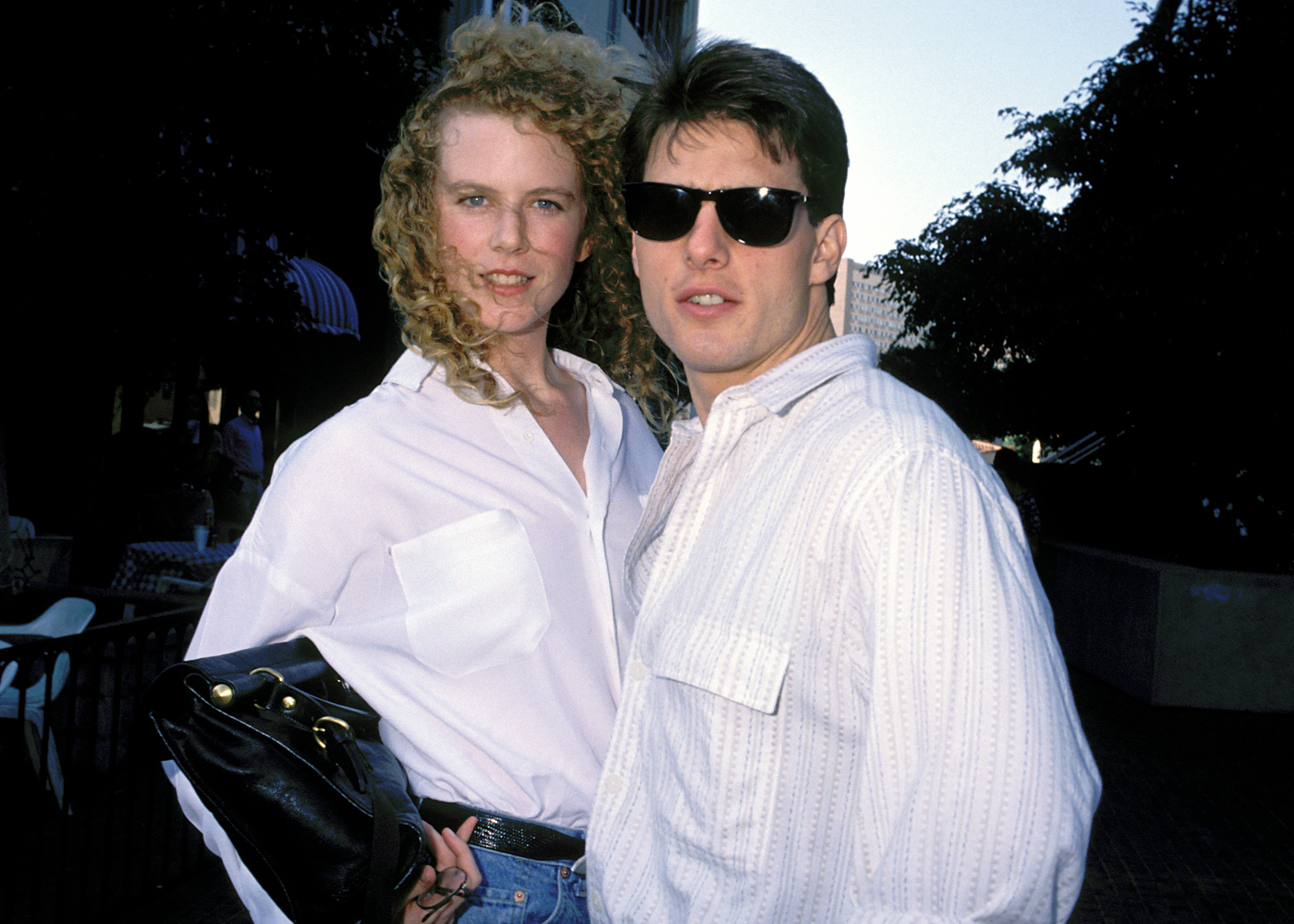 A woman with curly strawberry-blonde hair and a white shirt stands beside a dark-haired man wearing black sunglasses and a white striped shirt.
