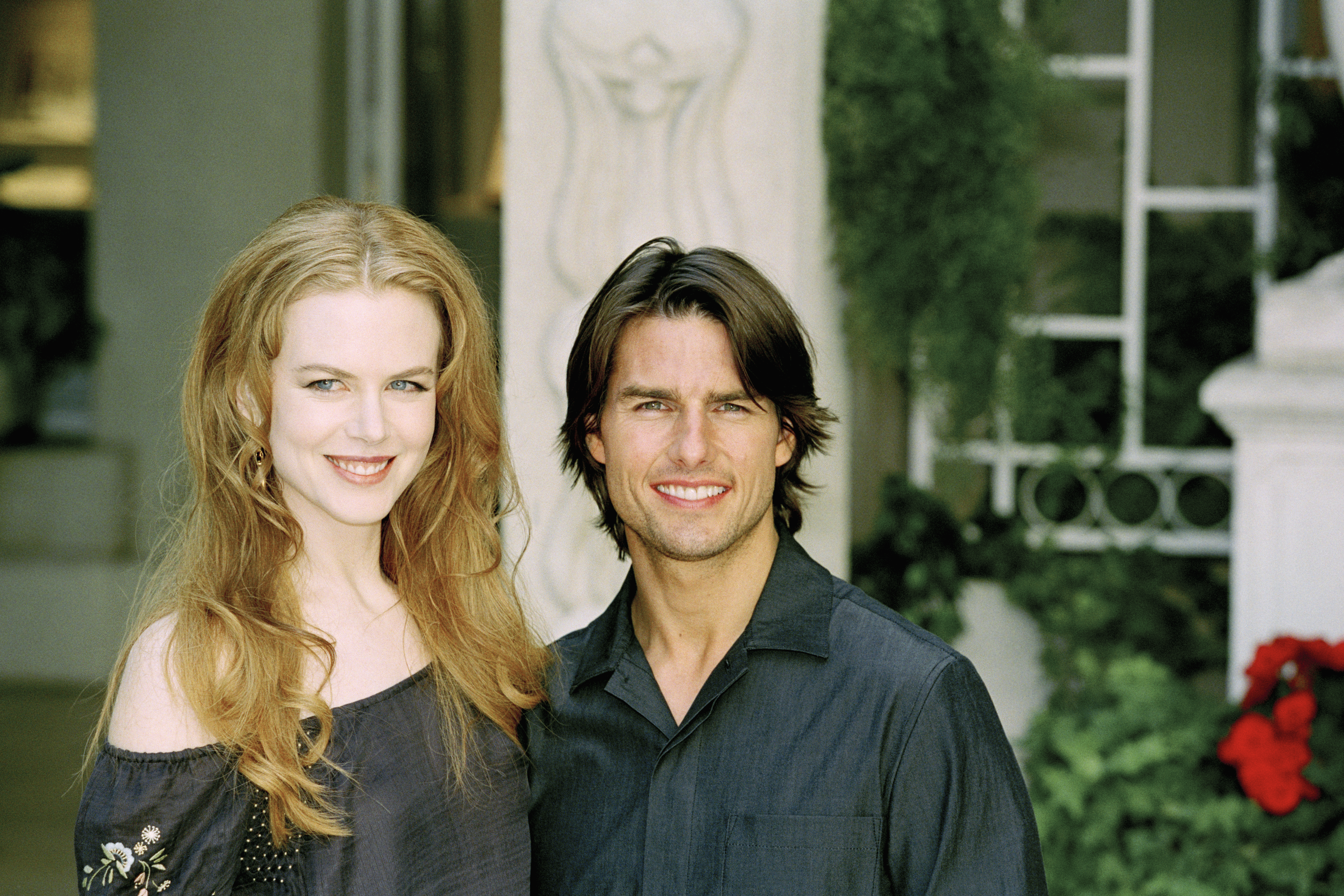 A woman with long red wavy hair in an off-shoulder top smiles next to a dark-haired man in a black shirt in front of a stone garden backdrop.