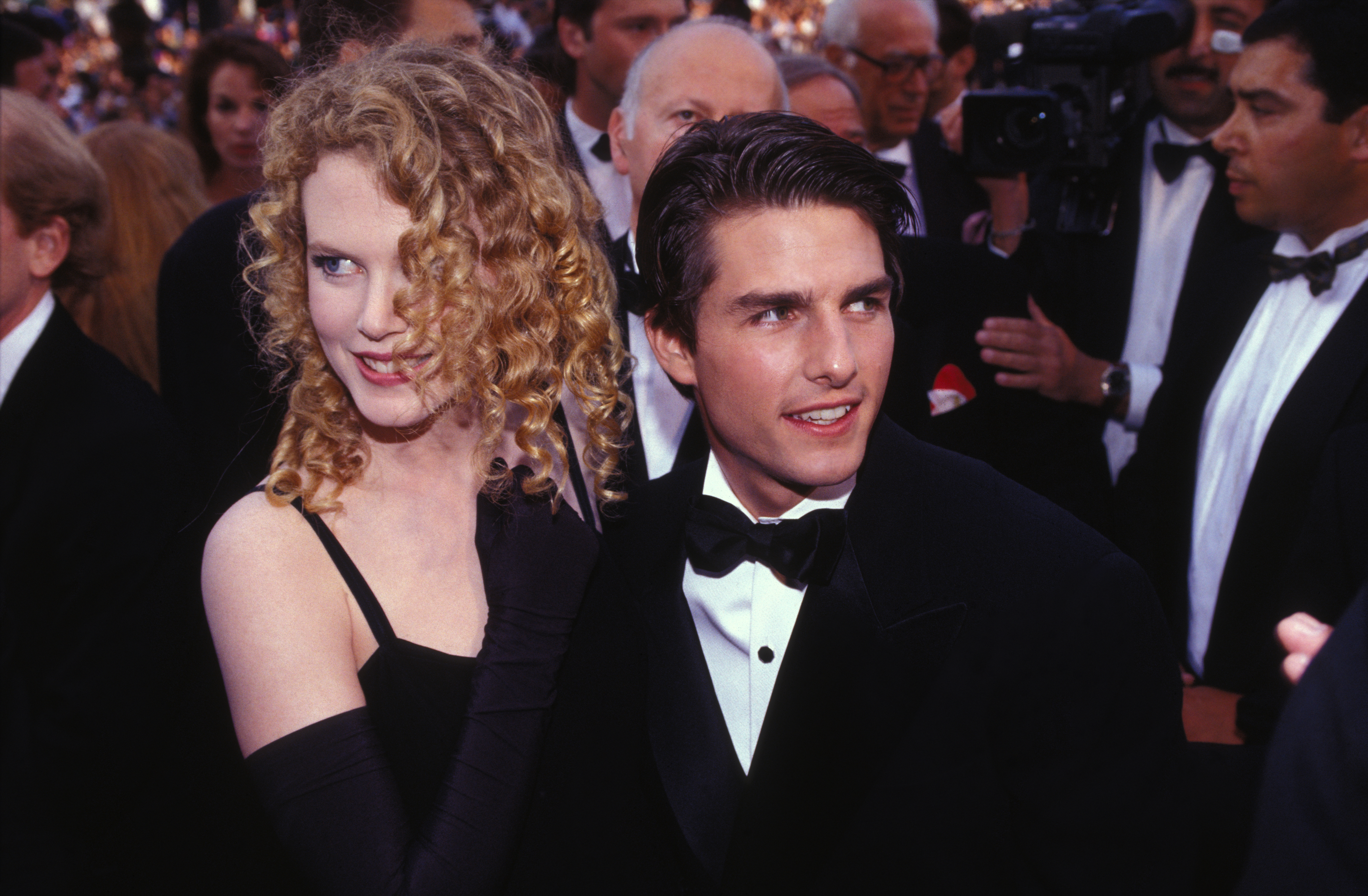 A curly-haired woman in a black gown stands beside a dark-haired man in a tuxedo amid a crowd at a formal event.
