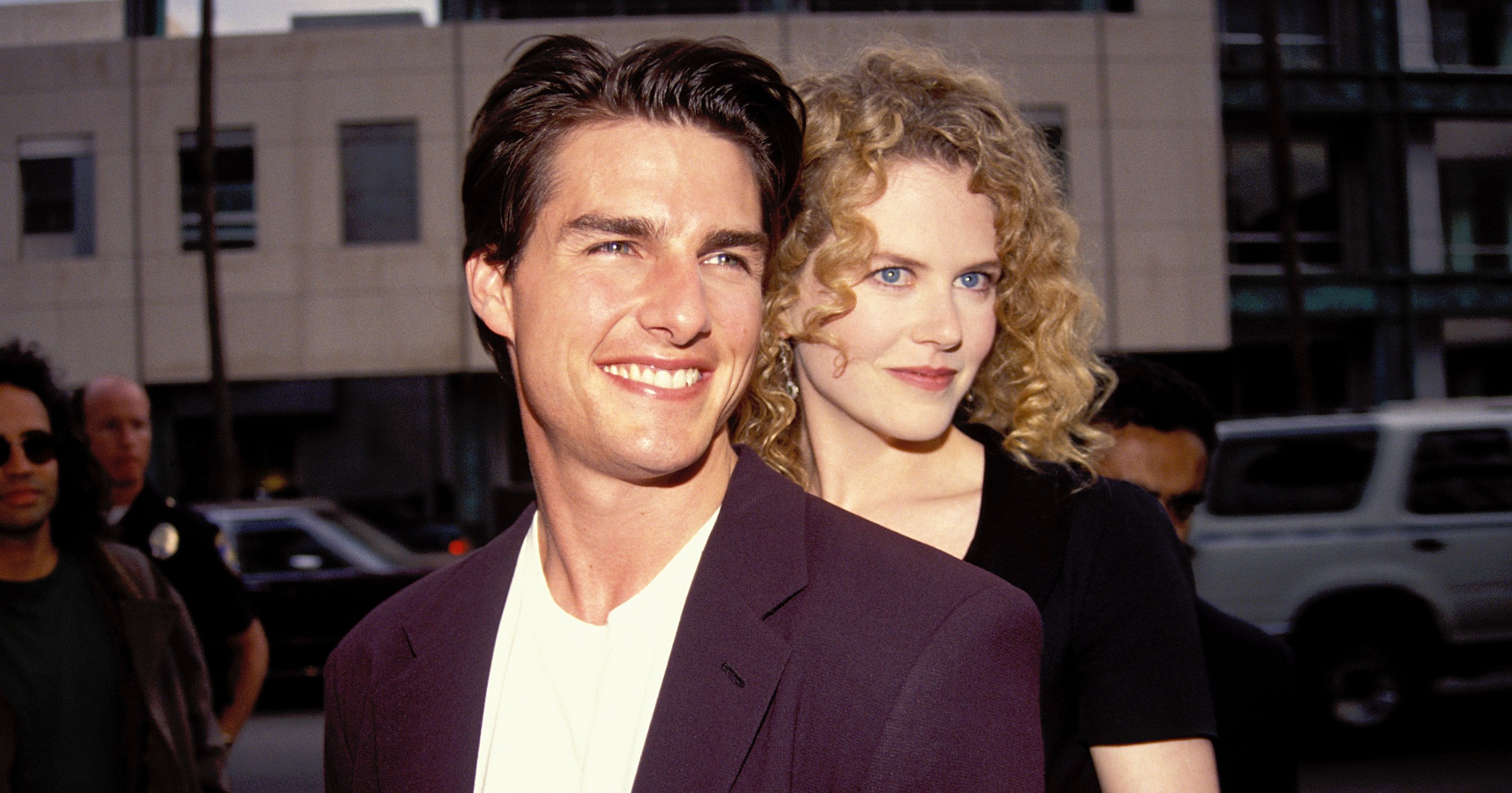 A smiling man in a dark blazer and white shirt stands in front of a curly-haired blonde woman who holds his arms from behind, outdoors in Los Angeles.