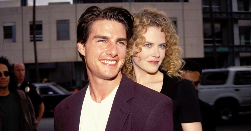A smiling man in a dark blazer and white shirt stands in front of a curly-haired blonde woman who holds his arms from behind, outdoors in Los Angeles.