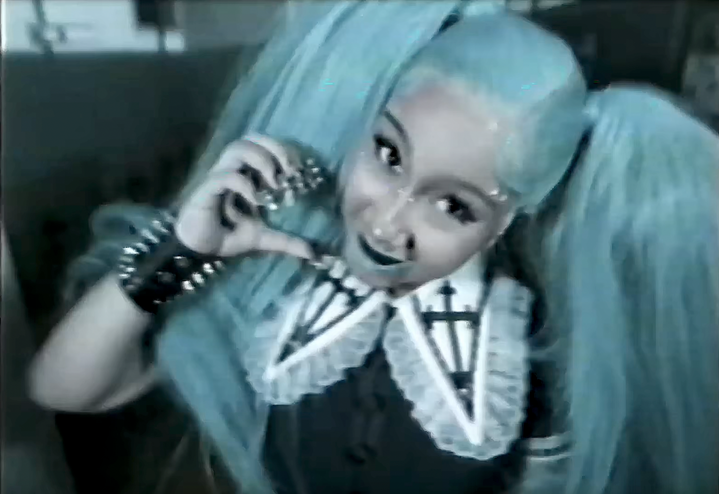 Person in gothic Lolita outfit with teal twin-tail wig, dark makeup, spiked bracelet, and cross necklace, posing against a dark background.