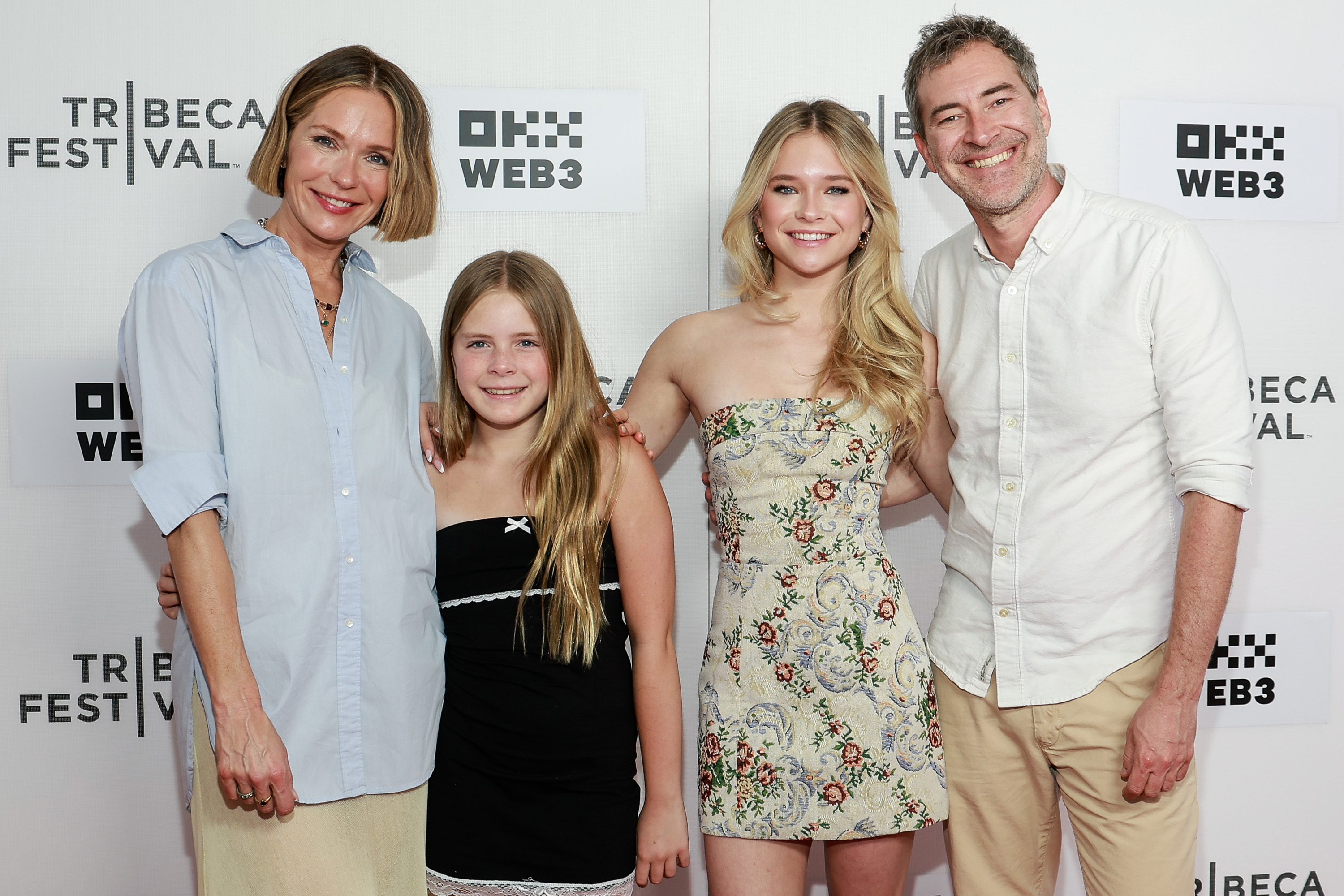 Four people smiling on a Tribeca Festival red carpet backdrop: a woman in a blue shirt, a young girl in black, a teen in a floral dress, and a man in a white shirt.