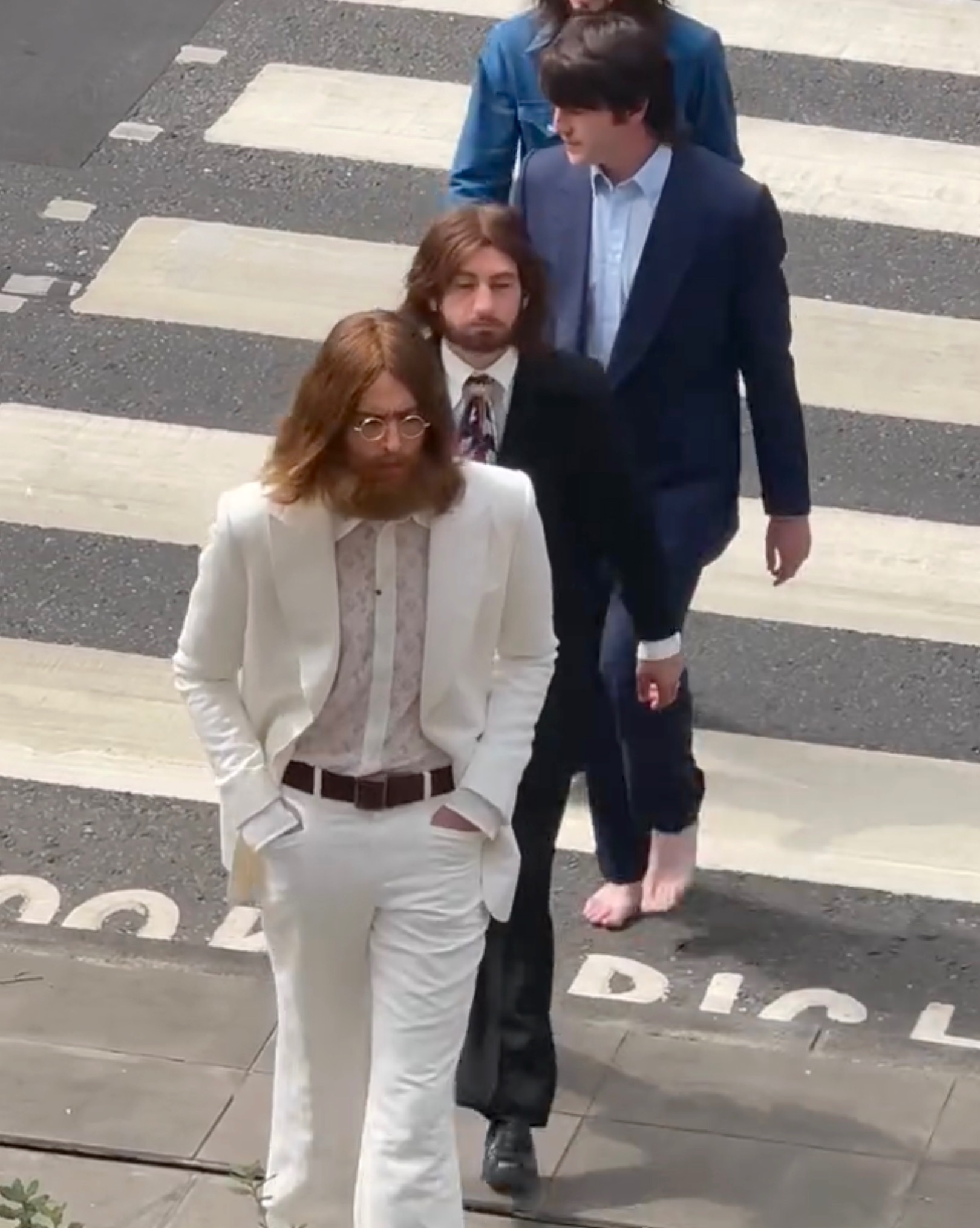 Close overhead view of four men on the Abbey Road crosswalk; the lead man wears a white suit with round glasses, one behind him is barefoot.