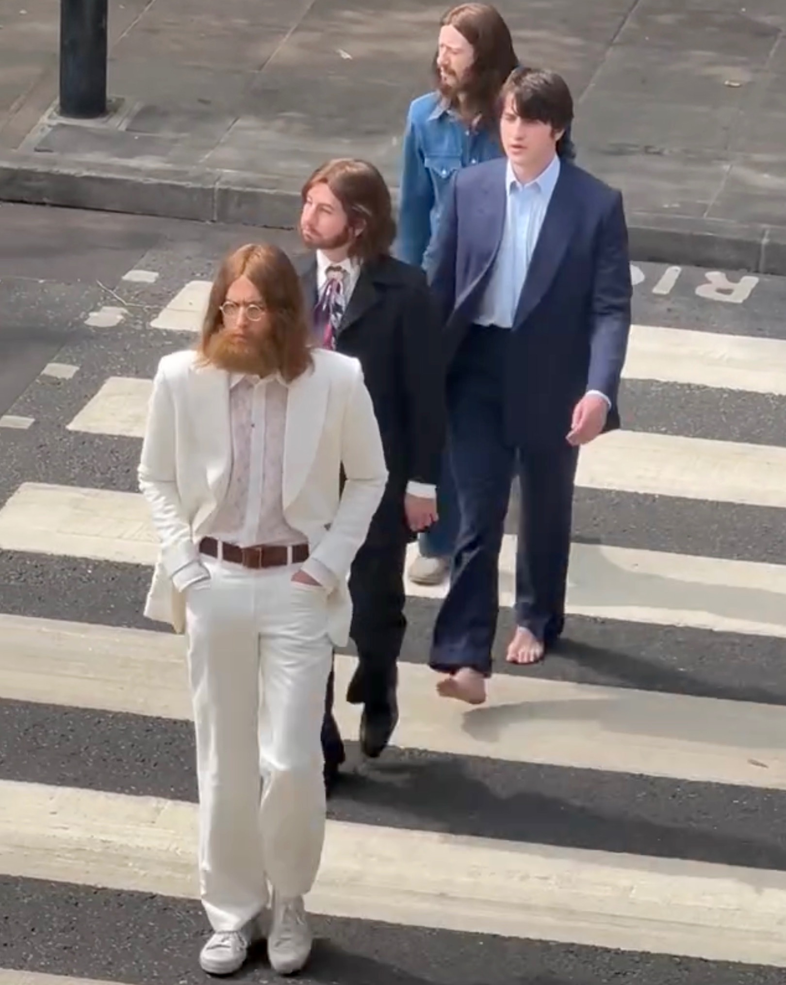 Four men walk across the Abbey Road zebra crossing; the man in front wears a white suit, the others in dark suits and a denim jacket.