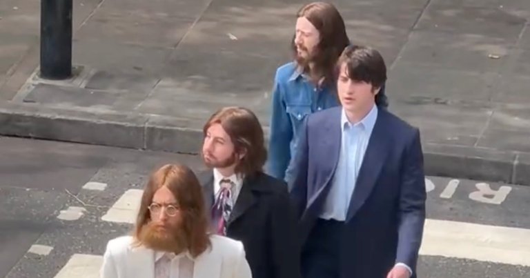 Four long-haired men in 1960s-style outfits — one in a white suit, one in black, one in a navy suit barefoot, one in denim — walk across the Abbey Road zebra crossing.
