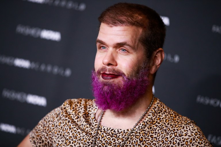 Man with a bright purple dyed beard and short brown hair wearing a leopard-print shirt and beaded necklaces at a red carpet event.