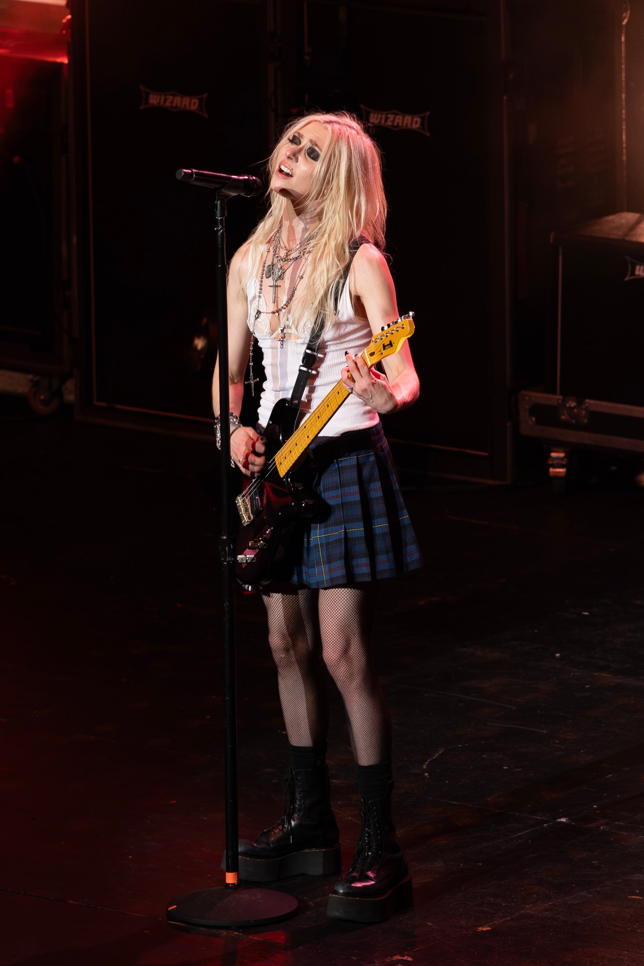 Taylor Momsen playing electric guitar and singing into a microphone on stage at The Wiltern, wearing a plaid skirt, white tank top, fishnet tights, and platform boots.