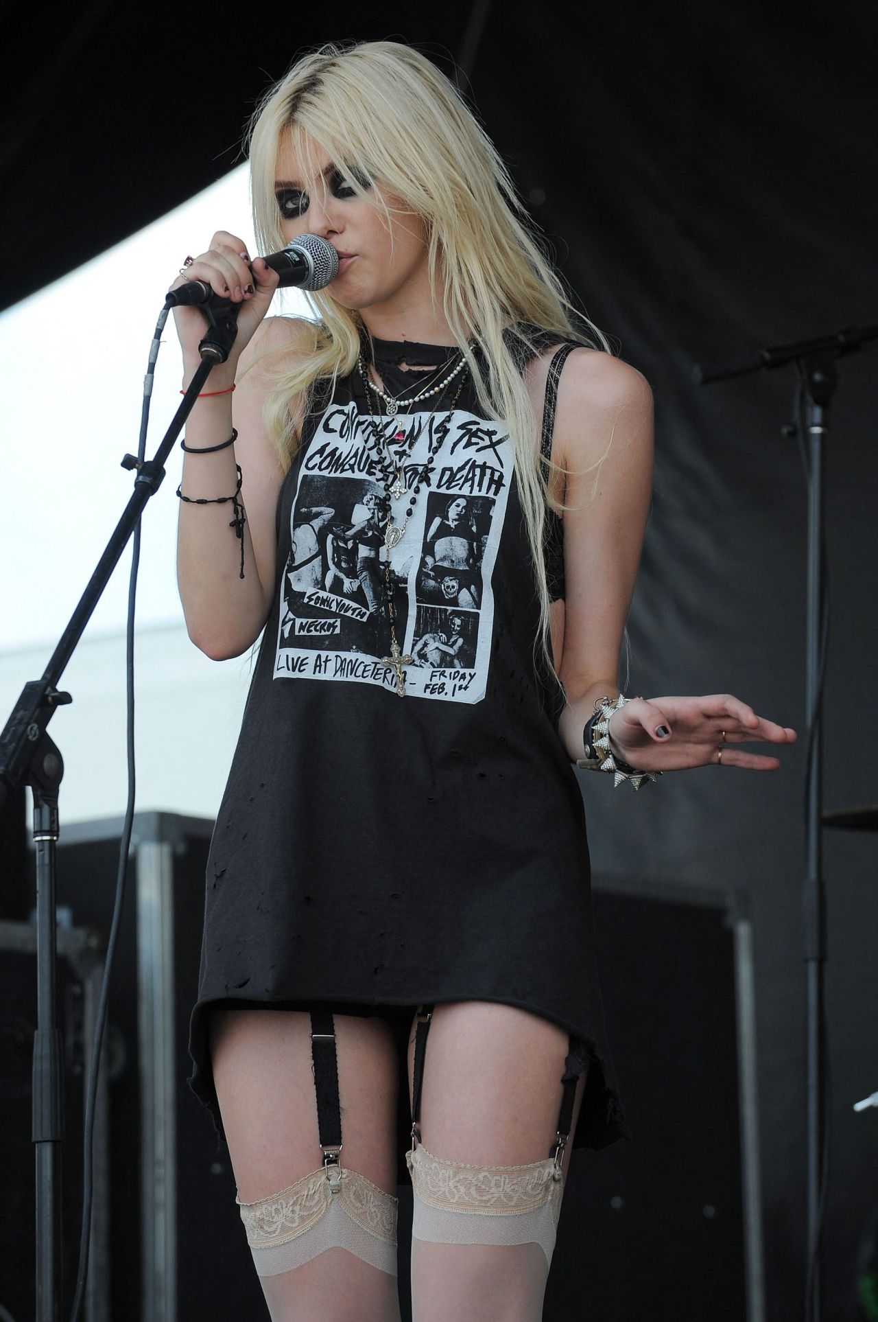 Teenage blonde singer performing on stage with a microphone, wearing a black graphic tee dress, garter belt, and lace stockings.