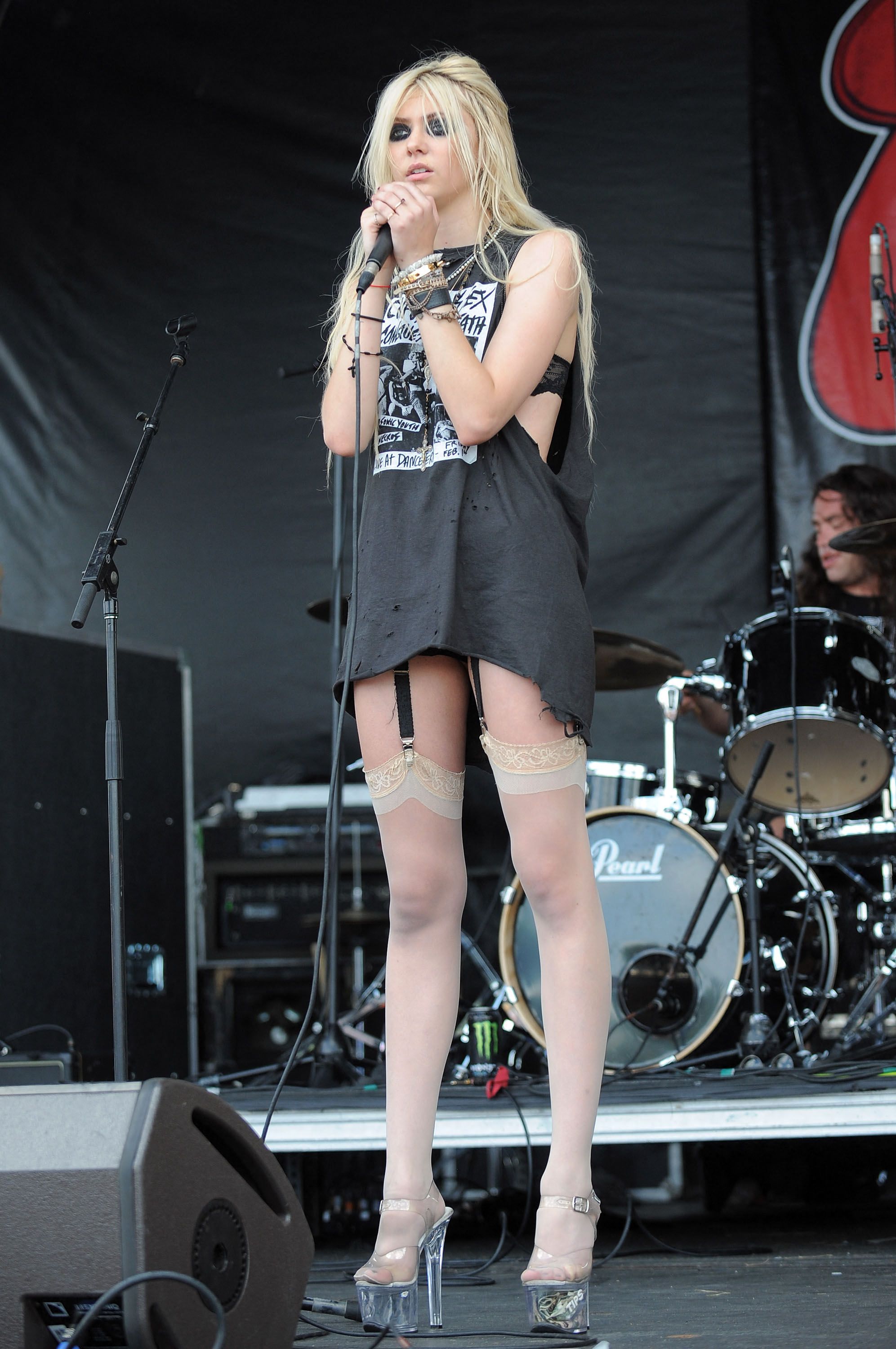 A teenage blonde female singer performs on an outdoor stage, holding a microphone, wearing a torn graphic tee, stockings with garters, and platform heels, with a drum kit behind her.