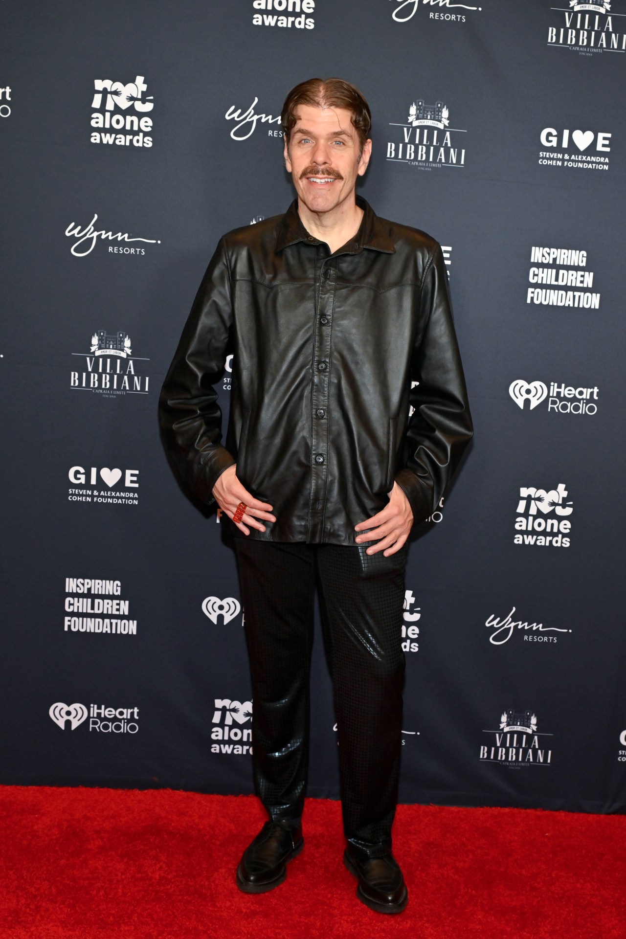 Perez Hilton wearing a black leather jacket and black pants, standing on a red carpet in front of a step-and-repeat banner at the Not Alone Awards.