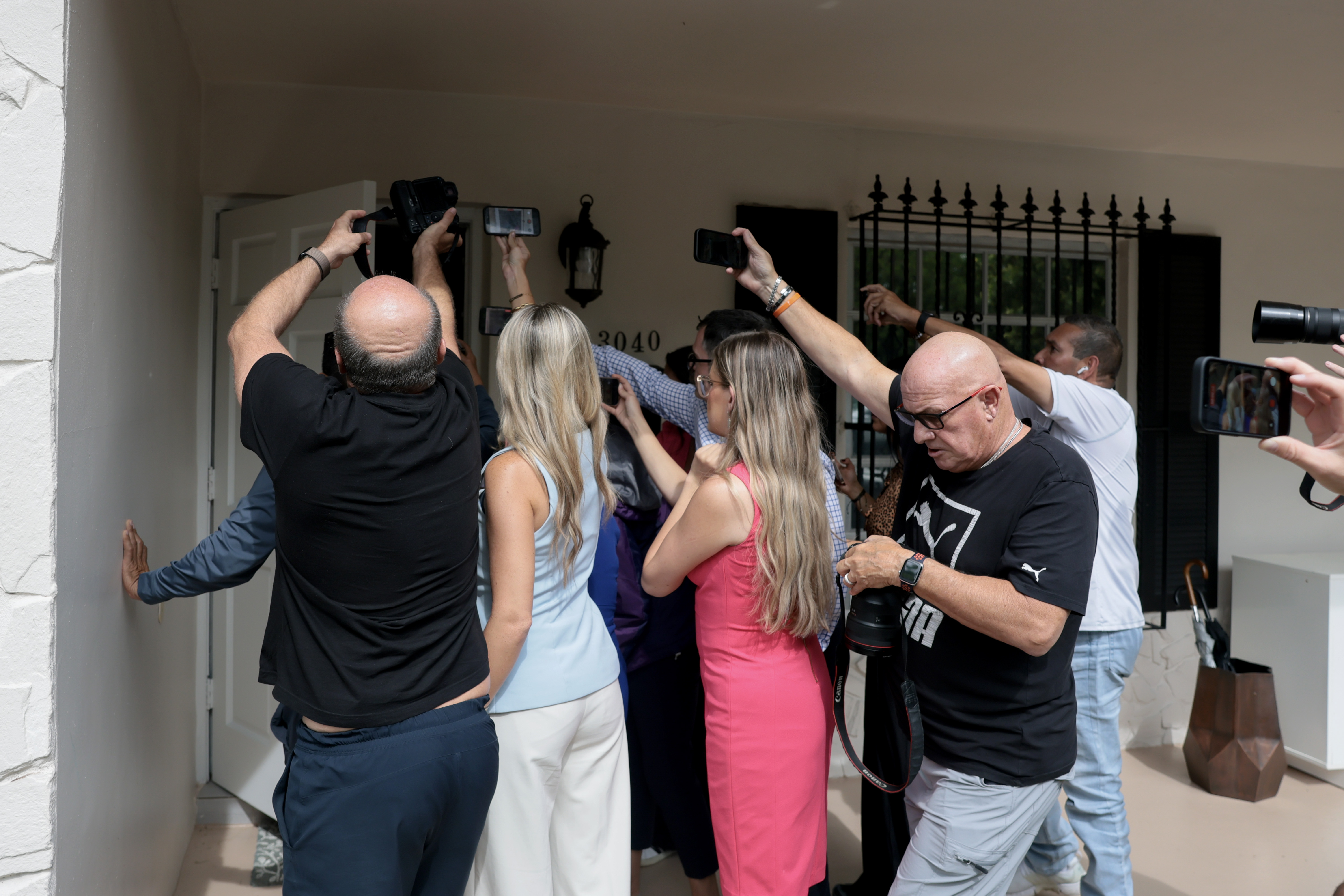 A crowd of journalists and photographers pressing toward the front door of a residential home numbered 3040 in Westchester, Florida.