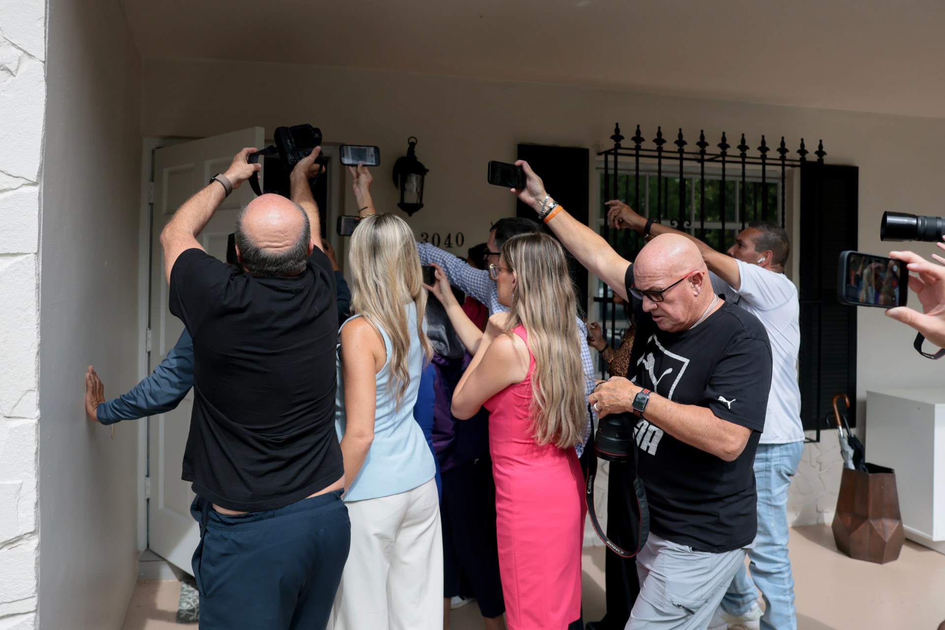 A crowd of journalists and photographers pressing toward the front door of a residential home numbered 3040 in Westchester, Florida.