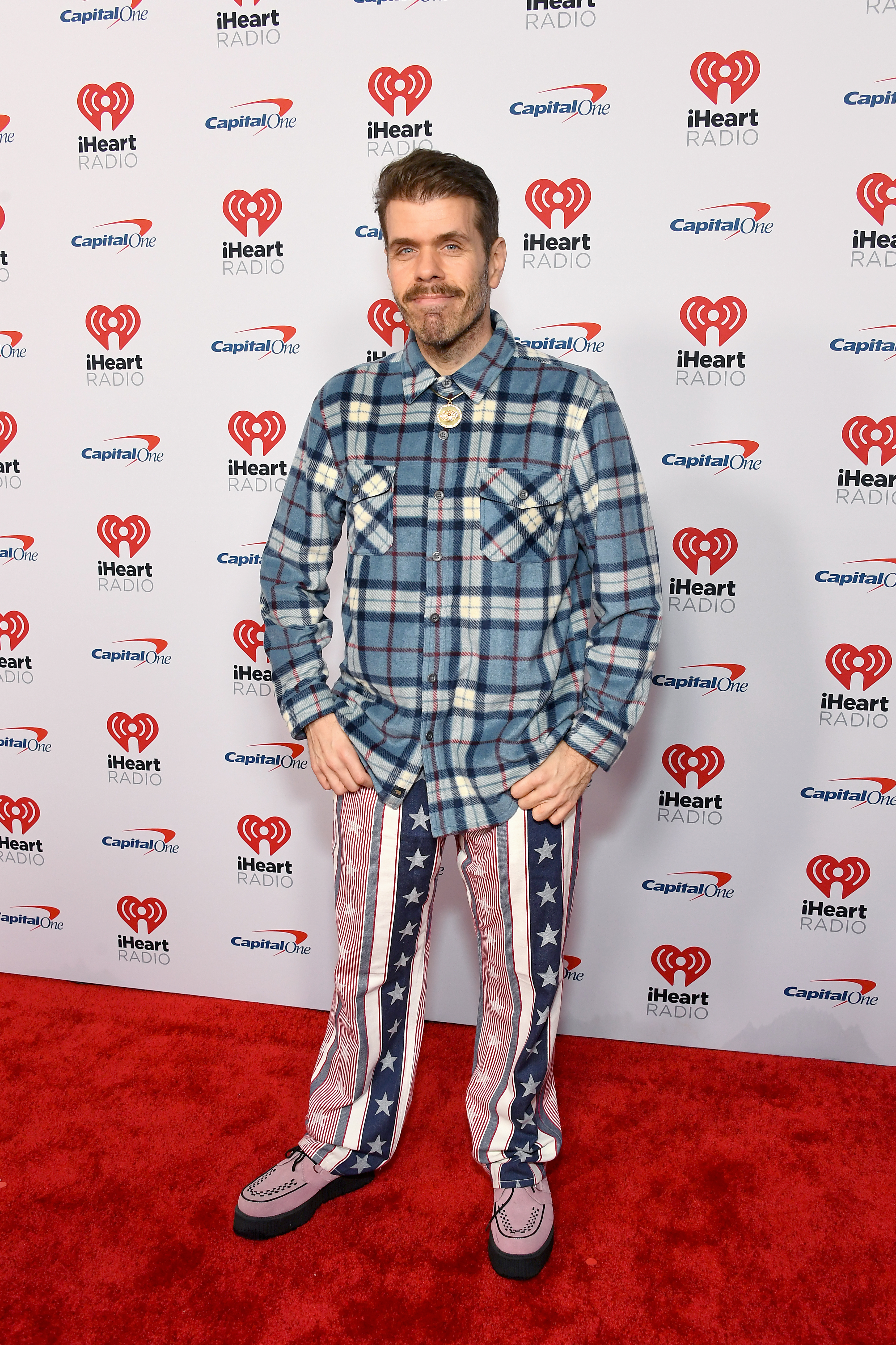 A man with a goatee wearing a blue plaid shirt and red, white, and blue star-and-stripes pants stands on a red carpet in front of an iHeartRadio and Capital One step-and-repeat backdrop.
