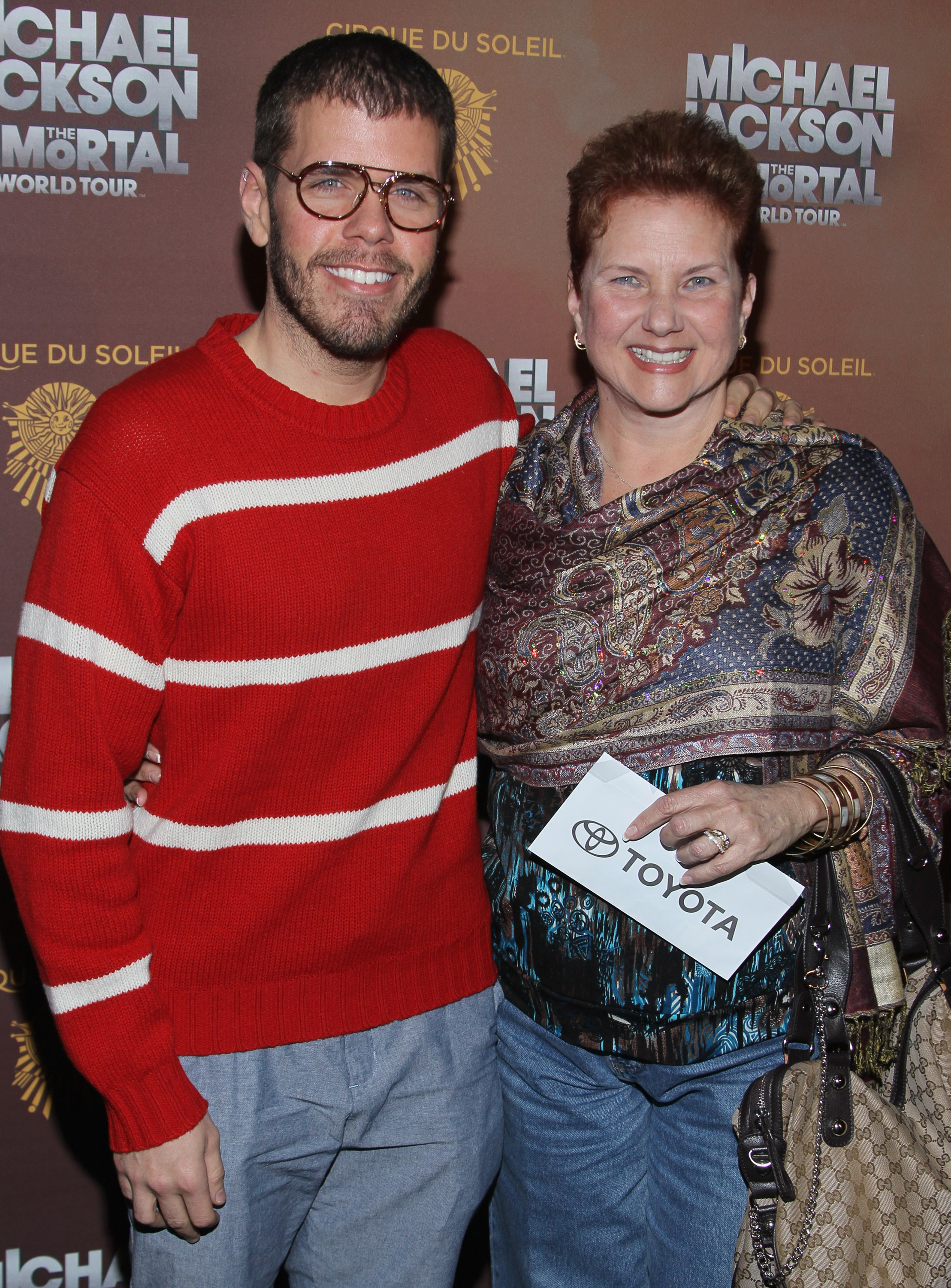 Perez Hilton in a red striped sweater and glasses stands smiling with his mother in front of a Michael Jackson Immortal World Tour step-and-repeat backdrop.