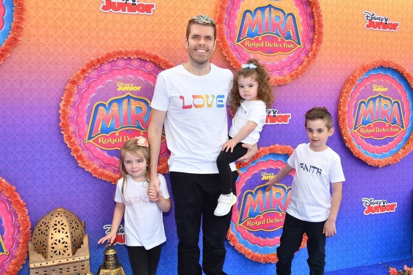 Man in a white 'LOVE' T-shirt holds a curly-haired toddler while standing with two other young children in front of a colorful Disney Junior 'Mira, Royal Detective' backdrop.