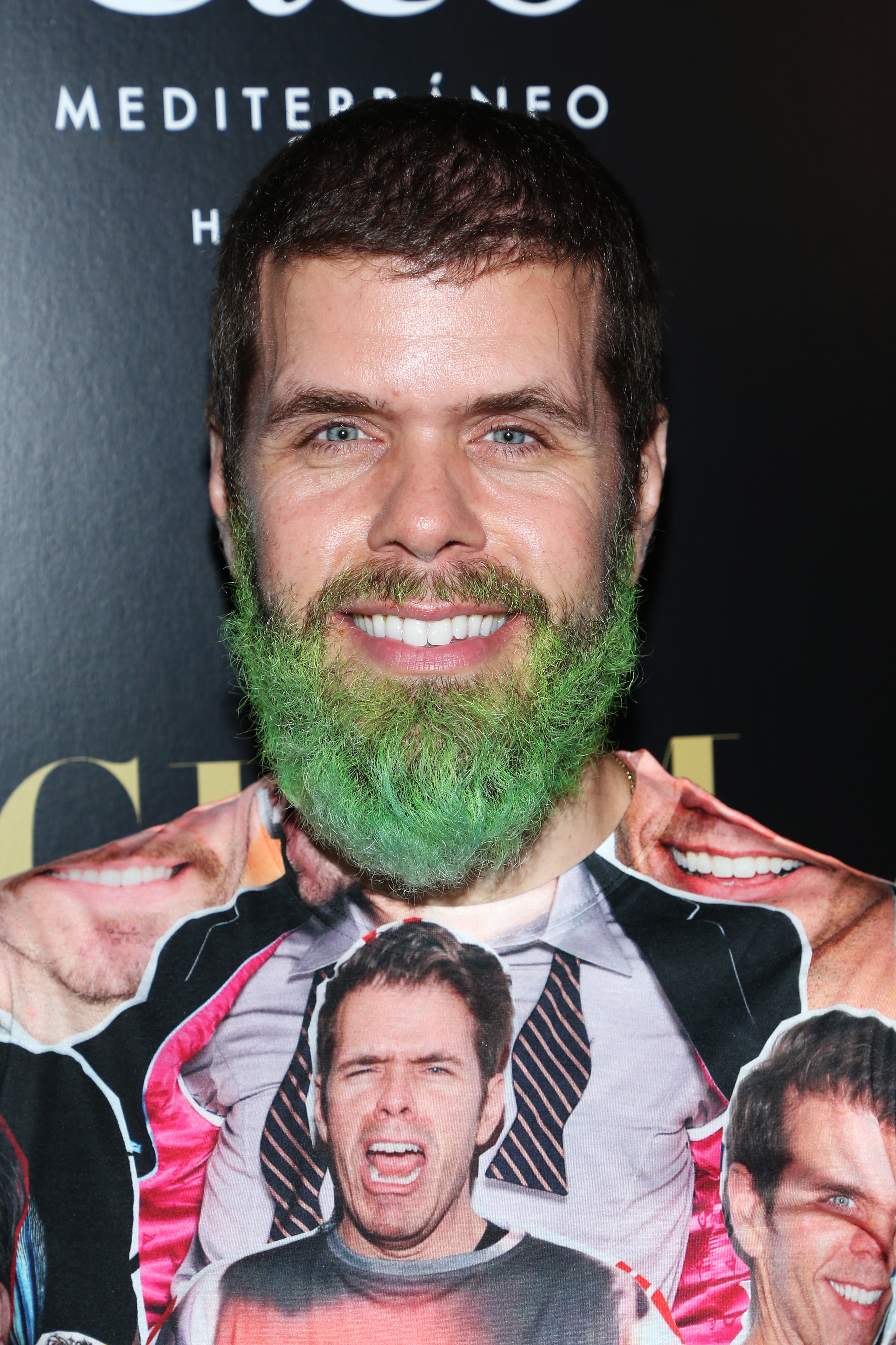 Man with a green-dyed beard and a novelty face-print shirt smiling at a Hollywood event in front of a dark branded backdrop.