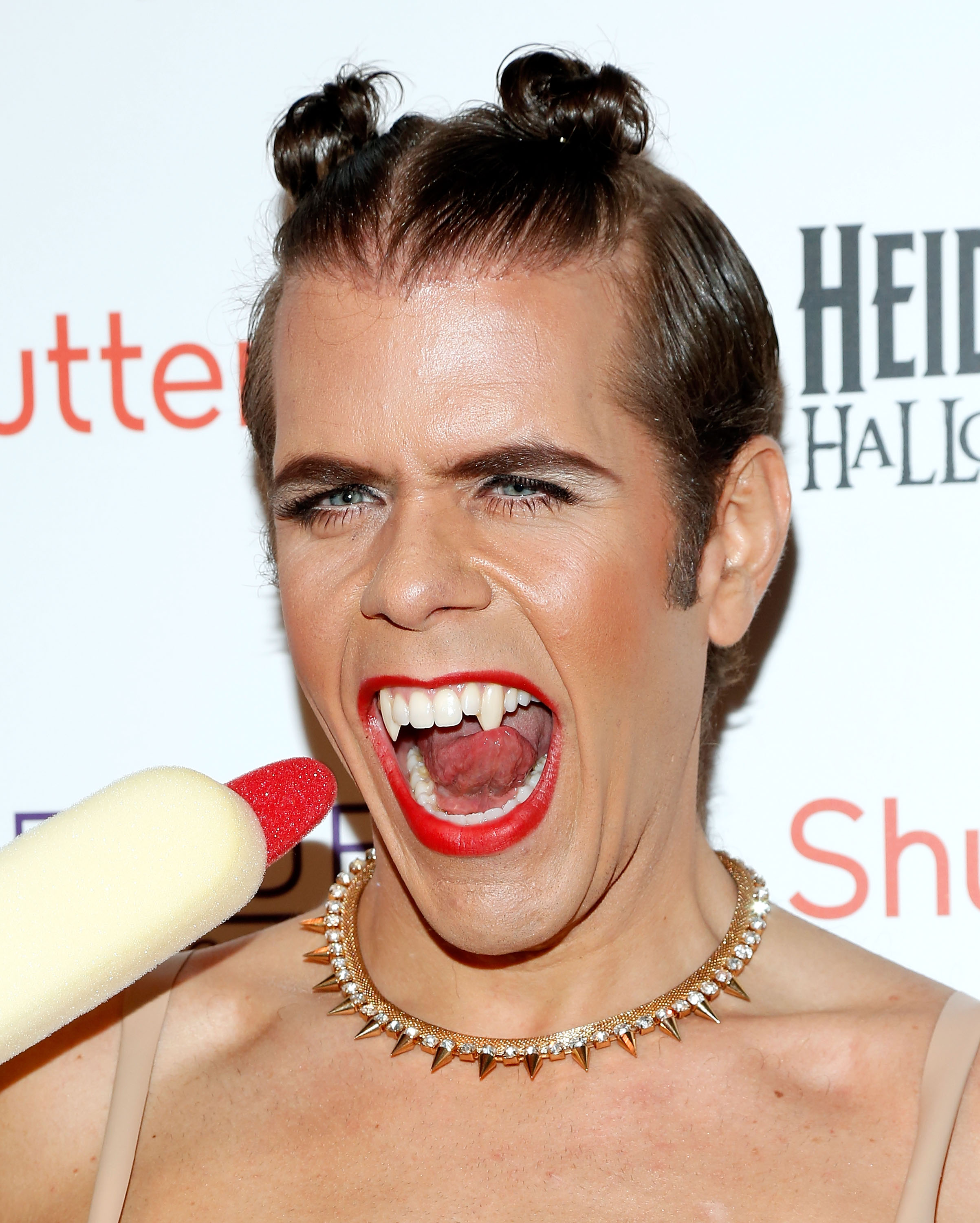 Person in vampire costume with red lipstick, fake fangs, double hair buns, and a spiked gold necklace, mouth wide open, holding a foam microphone at a Halloween party.
