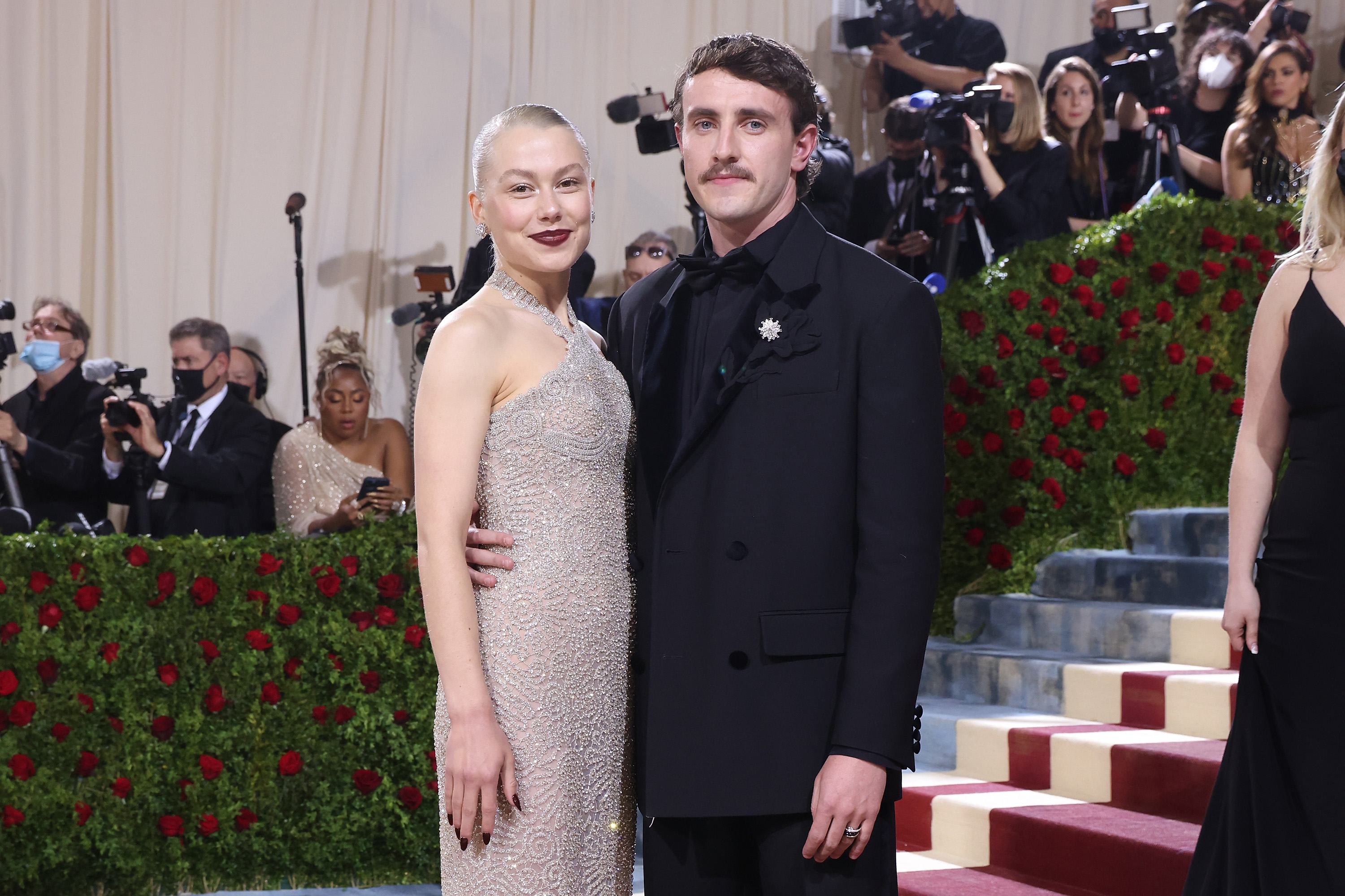 A woman in a beaded halter gown and a man in a black tuxedo with a brooch pose together on the Met Gala red carpet steps.