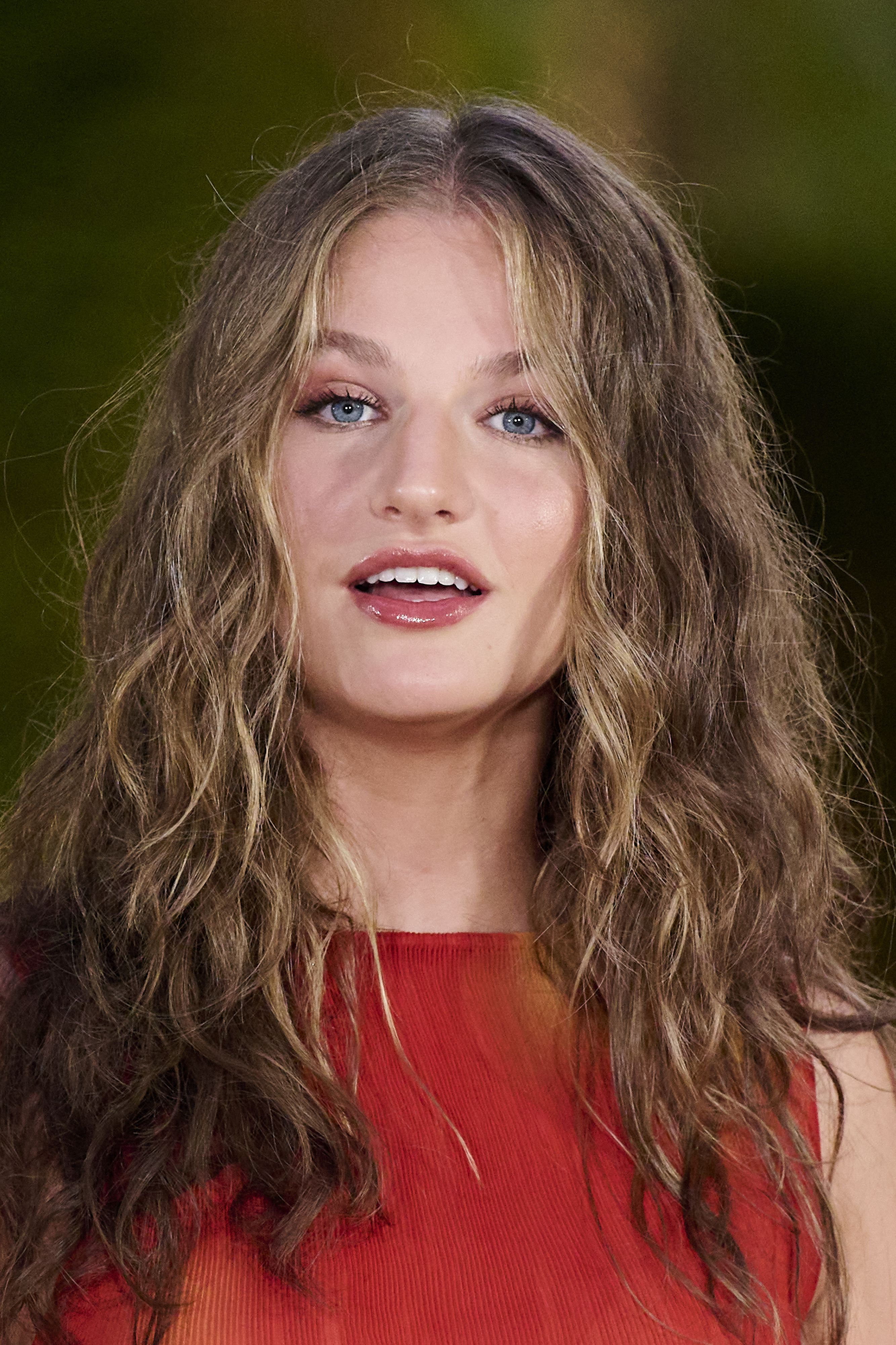 Close-up of Crown Princess Leonor of Spain with long, wavy brunette hair and a red sleeveless top, smiling slightly.