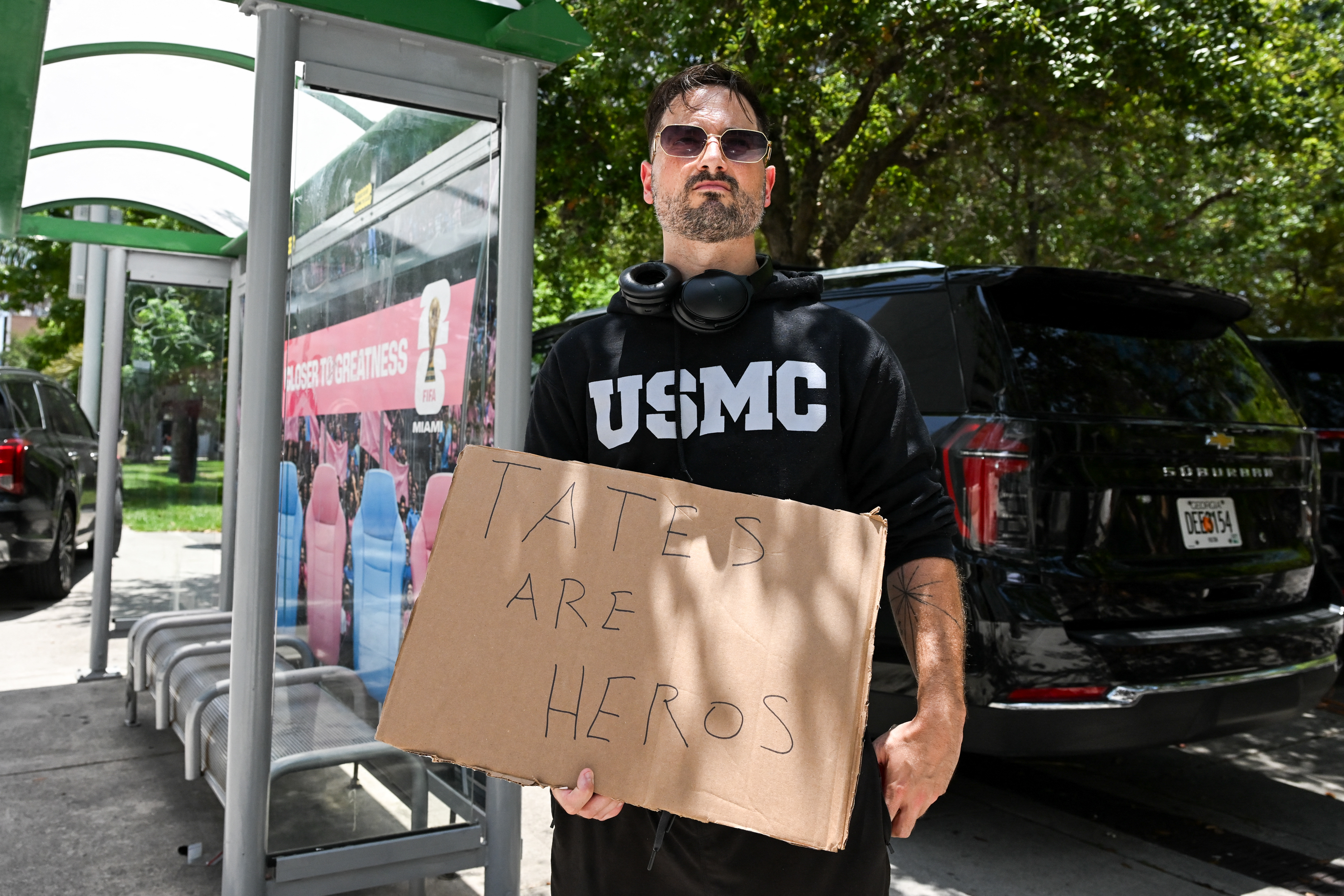 Man in a USMC hoodie and sunglasses holding a cardboard sign reading 'Tates Are Heros' at a bus stop outside a Miami courthouse.