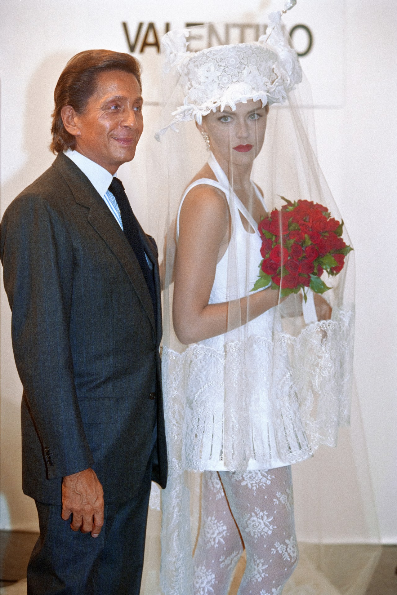 A man in a dark suit stands next to a woman dressed as a bride in a white lace gown and tall lace hat, holding a bouquet of red roses, in front of a Valentino sign.