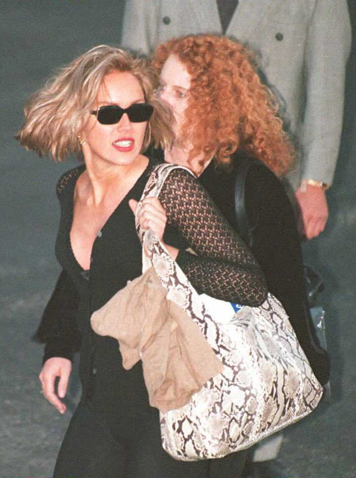 Blonde woman in black outfit and dark sunglasses walks through an airport alongside a woman with curly red hair carrying a snakeskin tote bag.