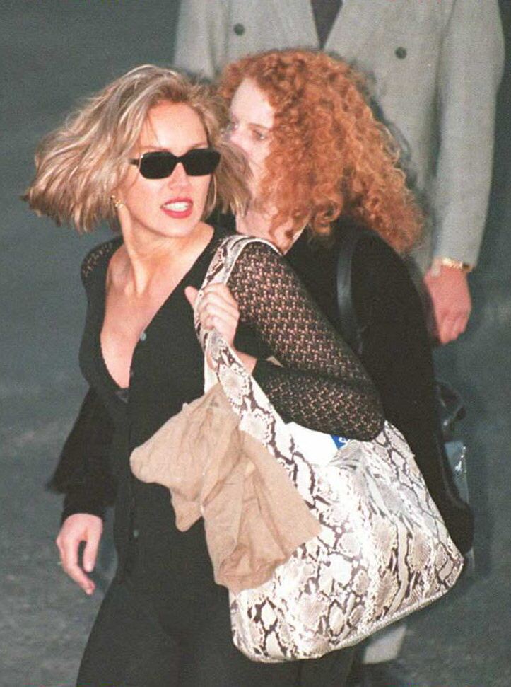 Blonde woman in black outfit and dark sunglasses walks through an airport alongside a woman with curly red hair carrying a snakeskin tote bag.