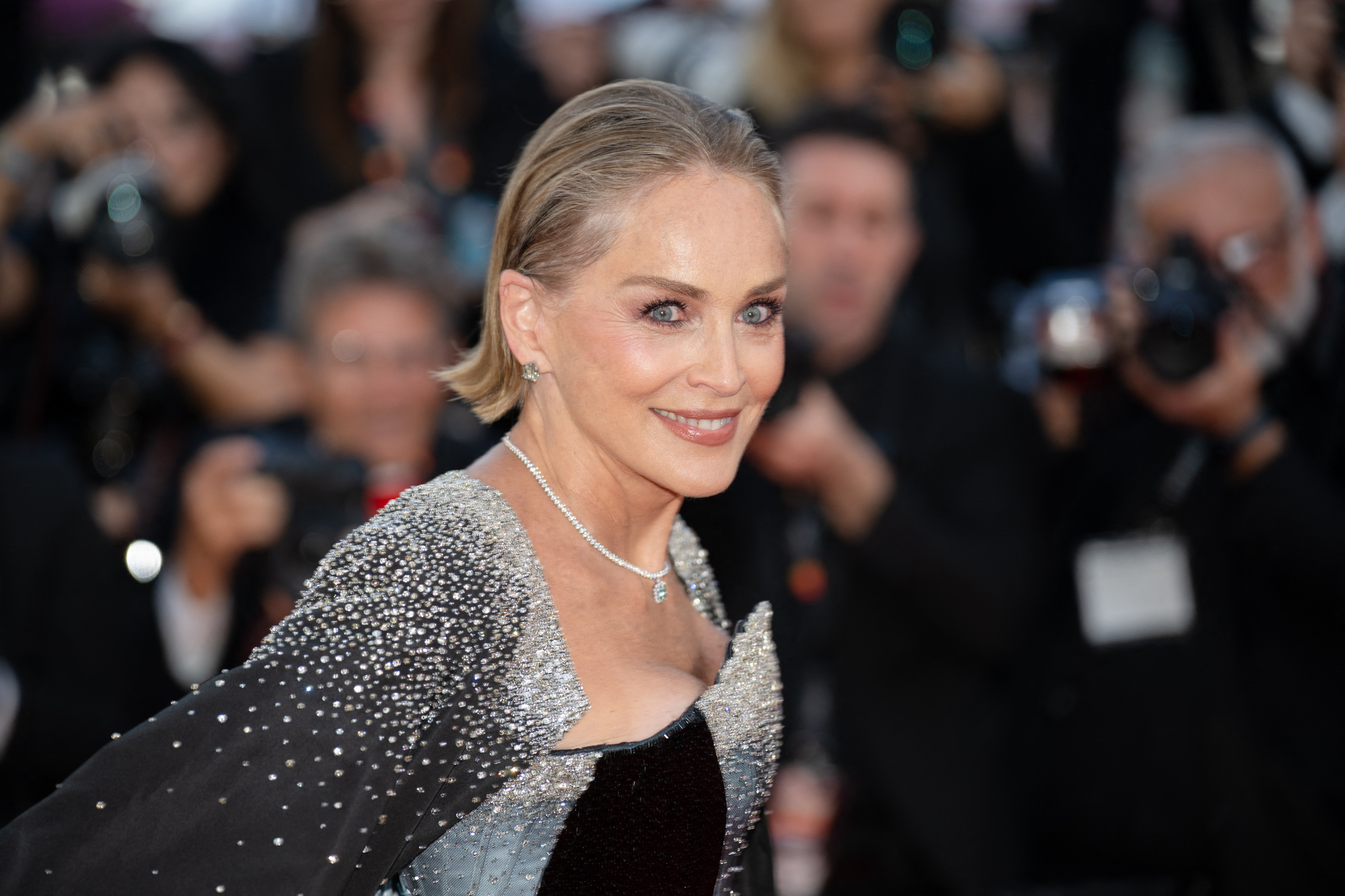 Woman with short blonde hair in a silver sequined cape and diamond necklace smiling over her shoulder at a red carpet event with photographers in the background.