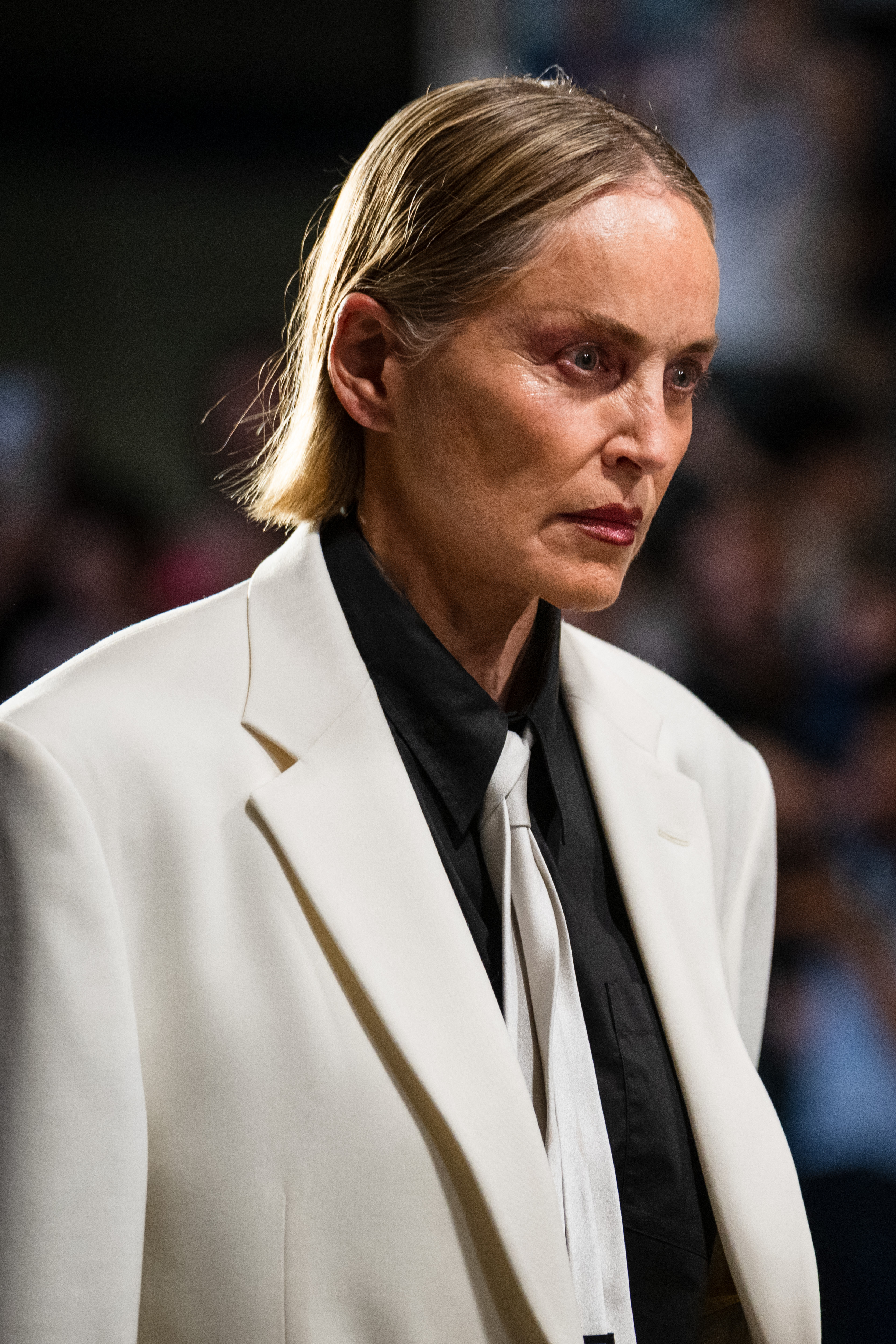 Sharon Stone on a runway in a white oversized blazer over a black shirt with a loose white tie, hair slicked back, looking intently forward.