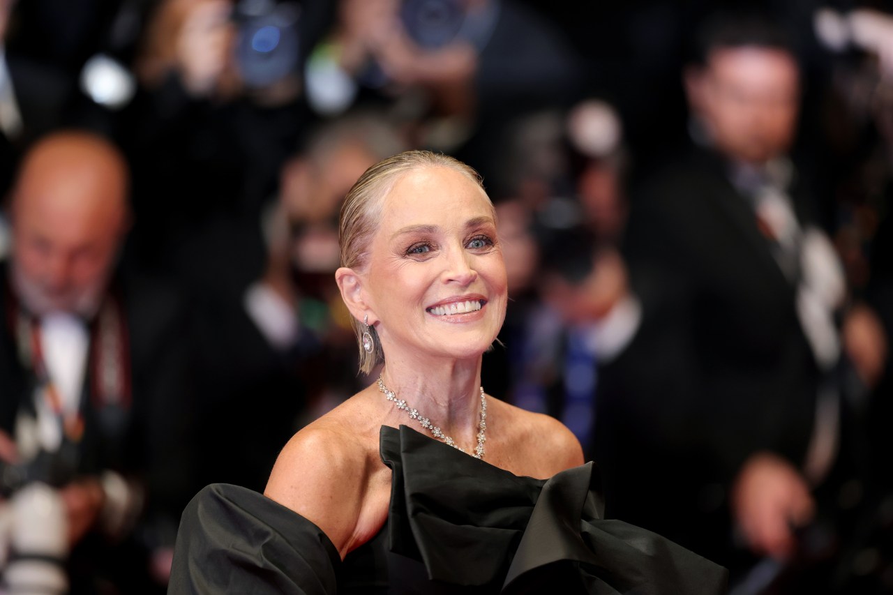 Woman in strapless black gown and diamond necklace smiling broadly at a red-carpet event, photographers blurred in background.