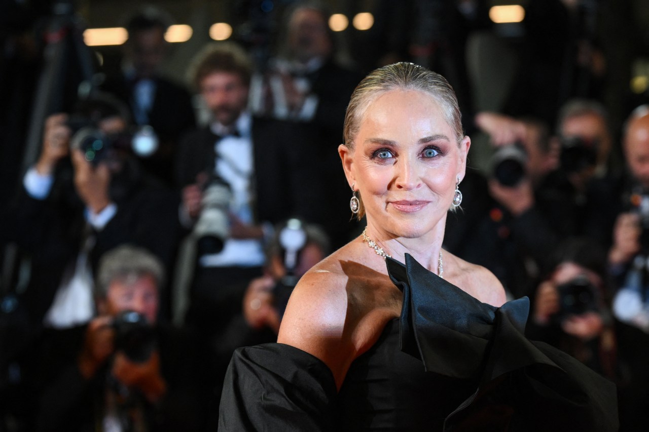Woman in off-shoulder black satin dress making a wide-eyed expression toward the camera at a film festival, photographers visible behind her.