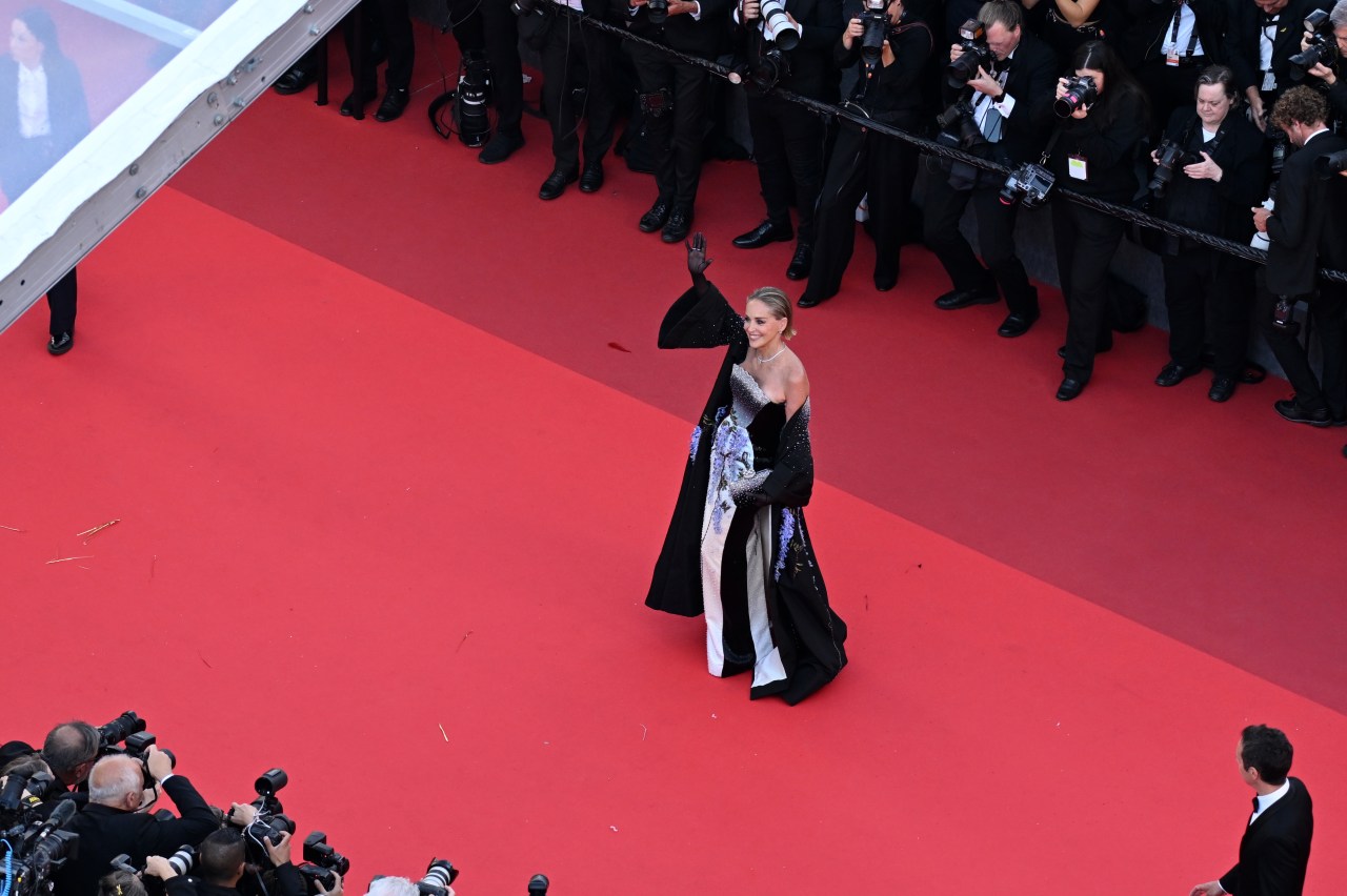 Aerial view of a woman in a floral gown and long black cape waving on a wide red carpet, surrounded by photographers along the edges.