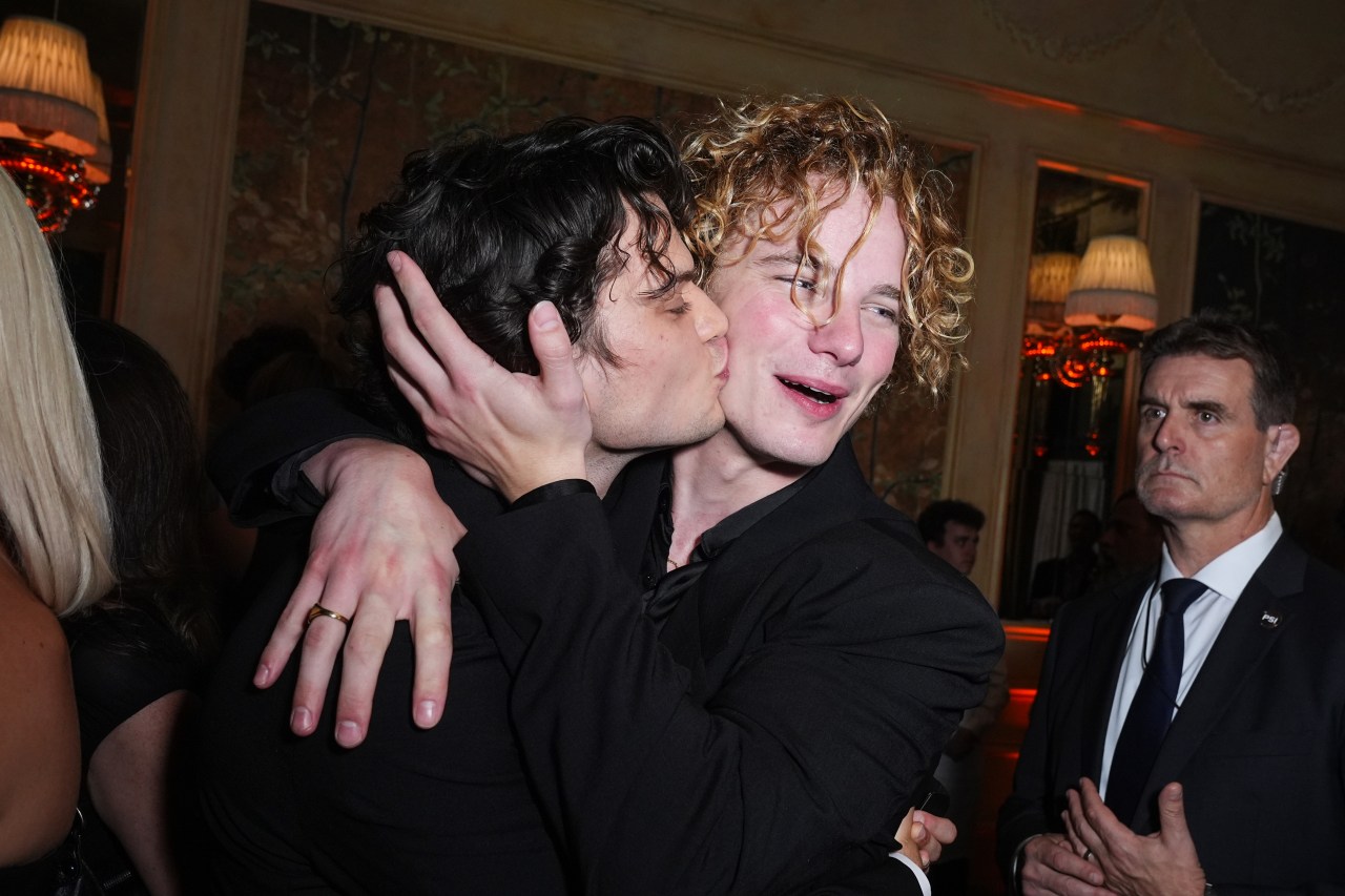 Homer Gere kissing Graham Campbell on the cheek as they embrace at an indoor evening party, both wearing black shirts.