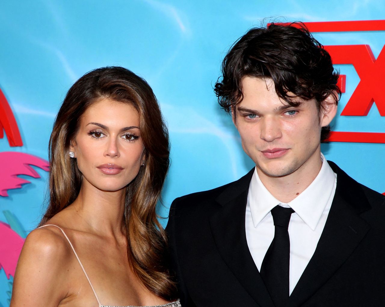 A young woman with long brown hair in a silver dress stands beside a dark-haired young man in a black suit and tie at a blue and red step-and-repeat backdrop.