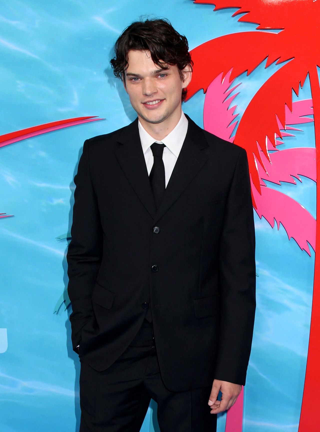 Young man with dark curly hair in a black suit and tie smiling at a blue and pink tropical-themed event backdrop.