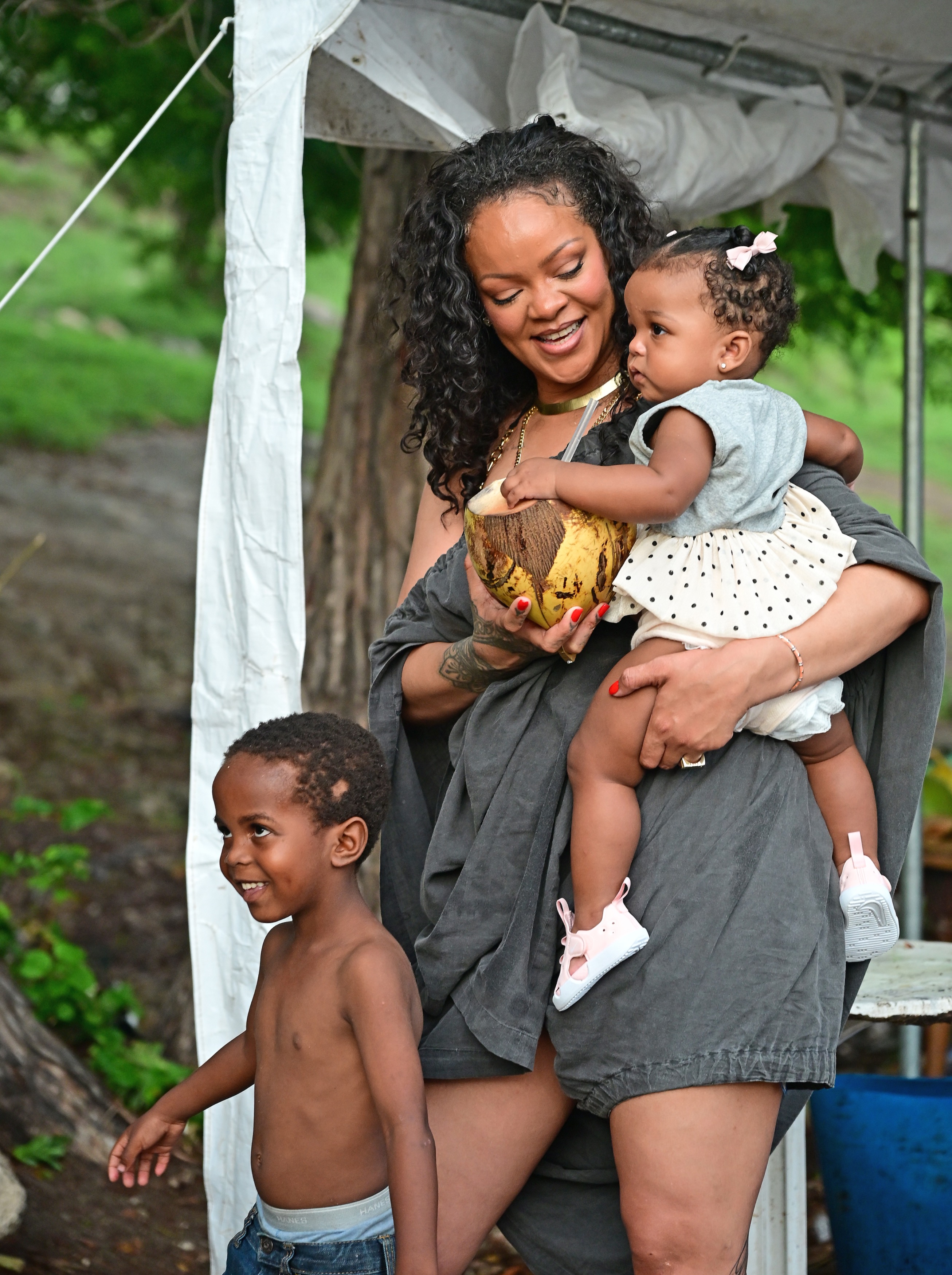A smiling woman in a gray outfit holds a baby girl in a polka-dot dress and a coconut, while a shirtless toddler boy stands nearby outdoors.