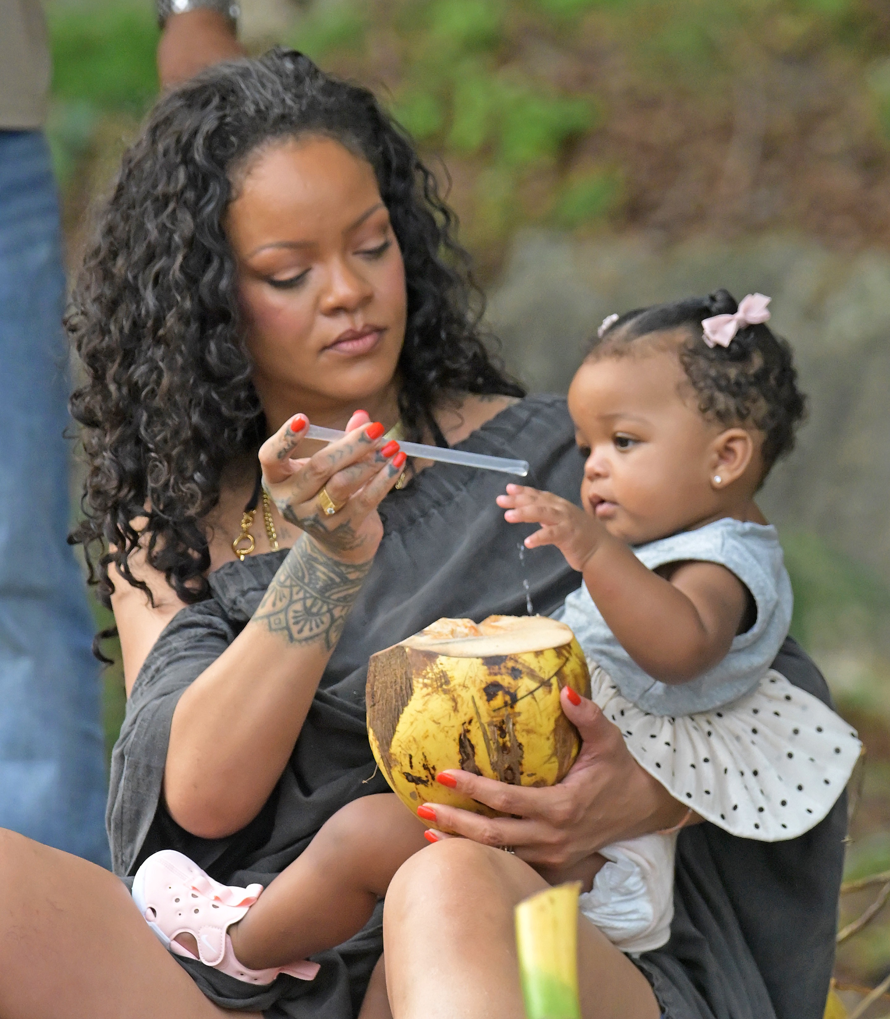 A curly-haired woman with red nails holds a coconut and a straw while a baby girl with a pink bow reaches toward the straw.