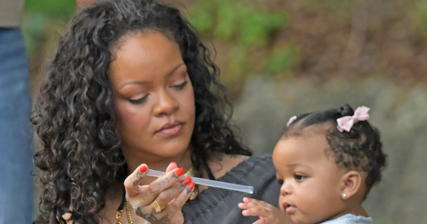 Woman with curly hair and hand tattoos holds a baby girl in a polka-dot dress while gripping an open coconut and a straw outdoors.