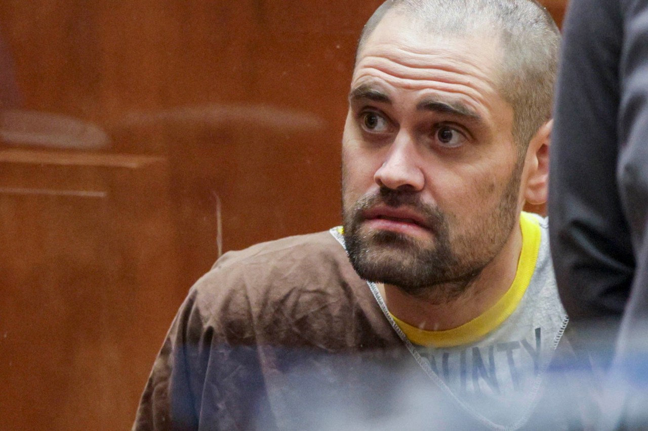 A man with a shaved head and short beard in a gray and yellow shirt sits in a courtroom, glancing sideways with a tense expression.