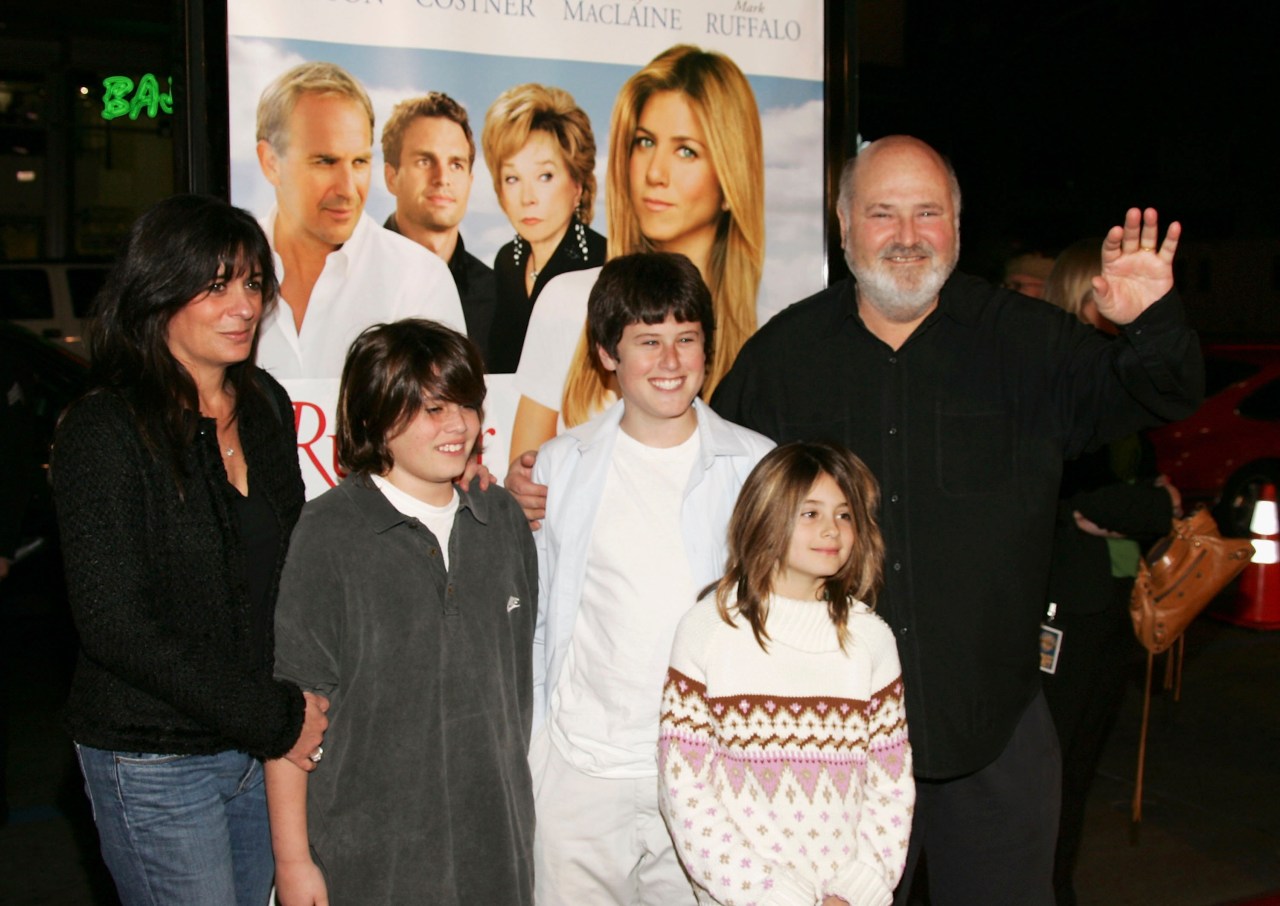 A man waving and a woman standing with three children in front of a movie premiere poster for 'Rumor Has It' outside a theater at night.