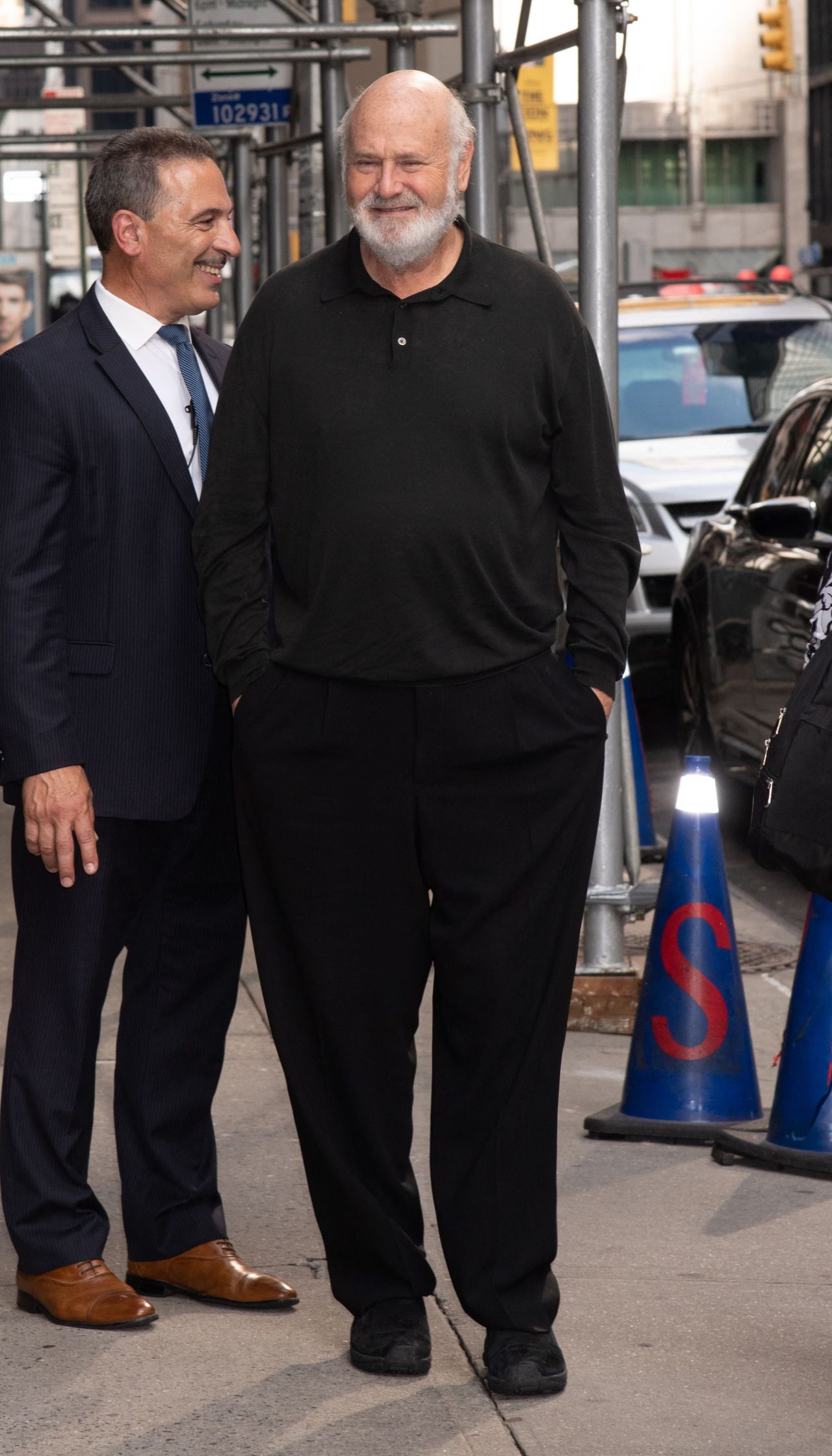Bald older man with white beard in all-black outfit walking on a NYC sidewalk next to a man in a navy suit, with traffic cones and parked cars behind them.