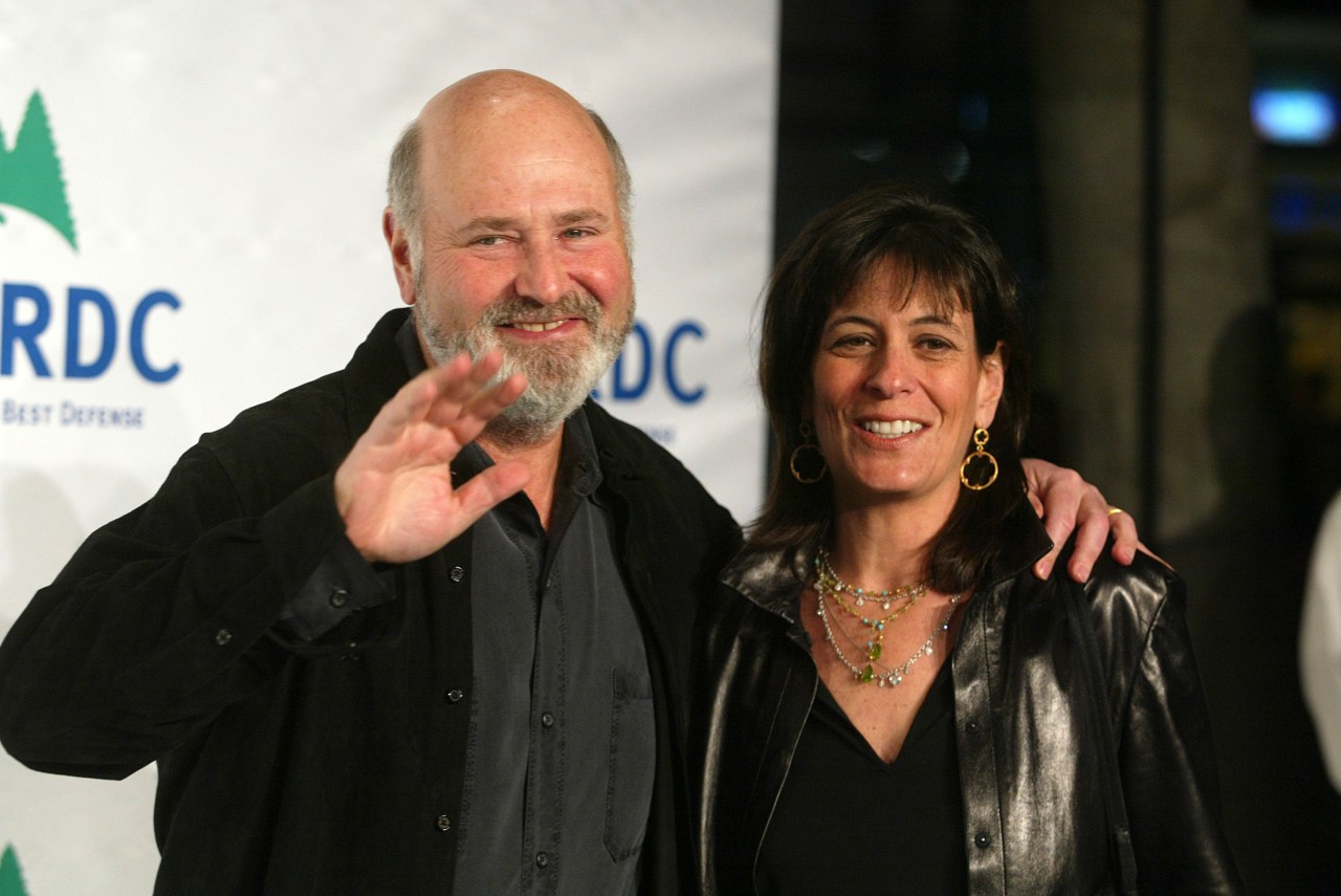 Bald man with gray beard waving and smiling with his arm around a dark-haired woman in a leather jacket, posed in front of an NRDC step-and-repeat backdrop.