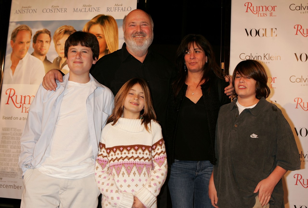 A bearded man in a black shirt stands with a woman and three children in front of a Rumor Has It movie premiere backdrop.